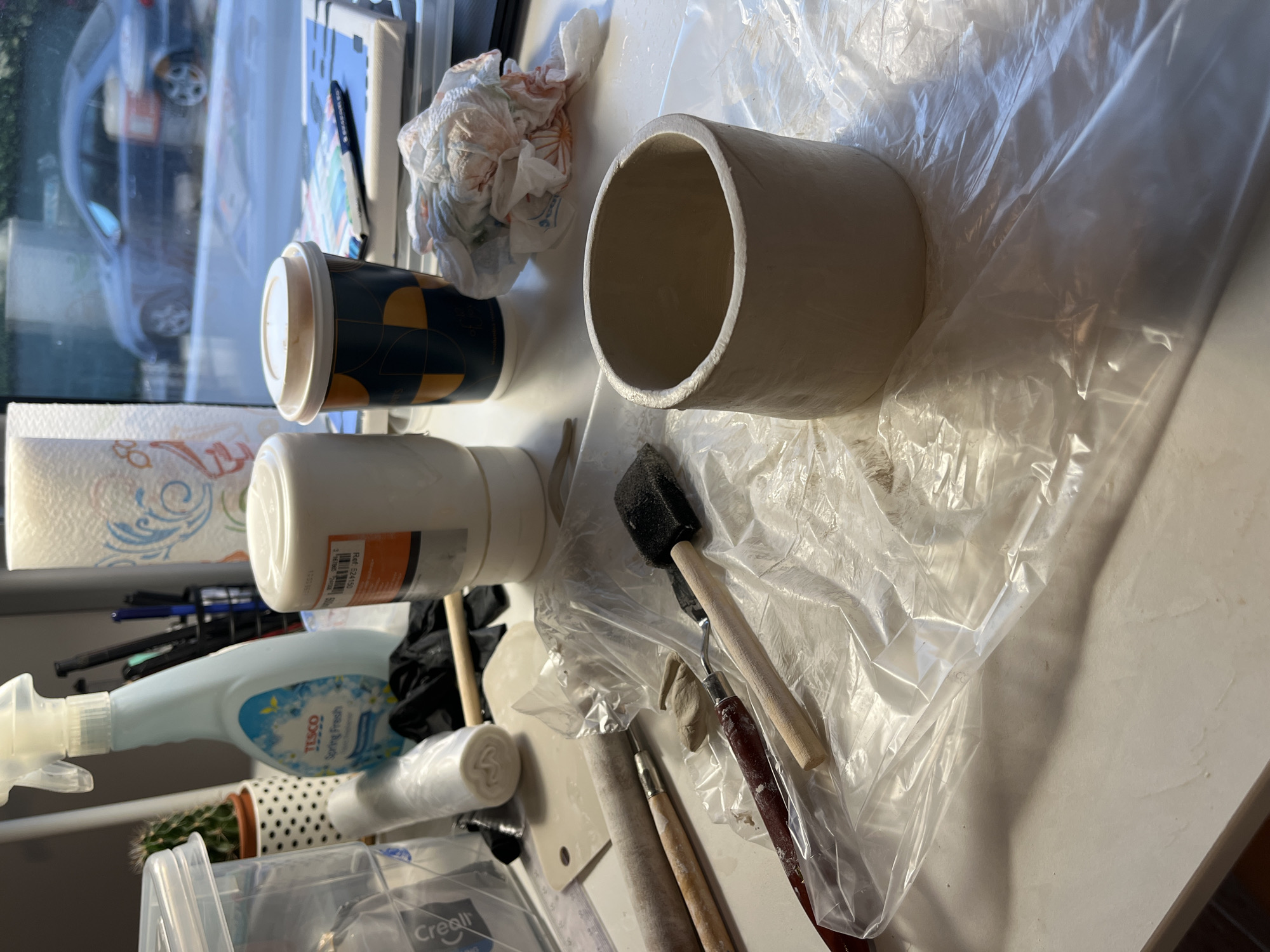 The image depicts a close-up, eye-level view of a cluttered workspace, likely a pottery or craft table. The primary subject is a cylindrical, unglazed clay pot, positioned in the foreground and center of the frame. It rests on a sheet of translucent plastic wrap that covers a portion of the table. Surrounding the pot are various crafting tools, including wooden-handled brushes with metal ferrules, one tipped with a black sponge, and another with what appears to be a rounded metal edge. A wooden dowel, possibly a rolling pin or a smoothing tool, is also visible, along with a small piece of clay.
In the mid-ground, to the left, is a blue spray bottle labeled "Tesco Spring Fresh". Next to it, a small potted cactus with spiky green leaves stands in a white pot with black polka dots. Further to the left, part of a clear plastic container is visible. Behind the spray bottle and cactus, a dark holder contains several pens and pencils. A roll of white material, possibly paper towels or more plastic wrap, is situated behind the crafting tools.
In the background, a white paper towel roll with a decorative blue and red pattern stands tall. To the right of this, a disposable coffee cup with a lid, featuring a dark blue and gold geometric pattern, is placed on the table. Adjacent to the coffee cup, a crumpled wad of paper towels with pink and orange stains is visible.
The scene appears to be indoors, with a window in the background. Through the window, a dark-colored car is visible, suggesting an urban environment. The lighting is soft, casting shadows across the table, indicating it could be daytime. The overall impression is one of active creative work in progress. The image does not provide any information that would allow for the specific determination of its location within the city of Limassol, Cyprus.
