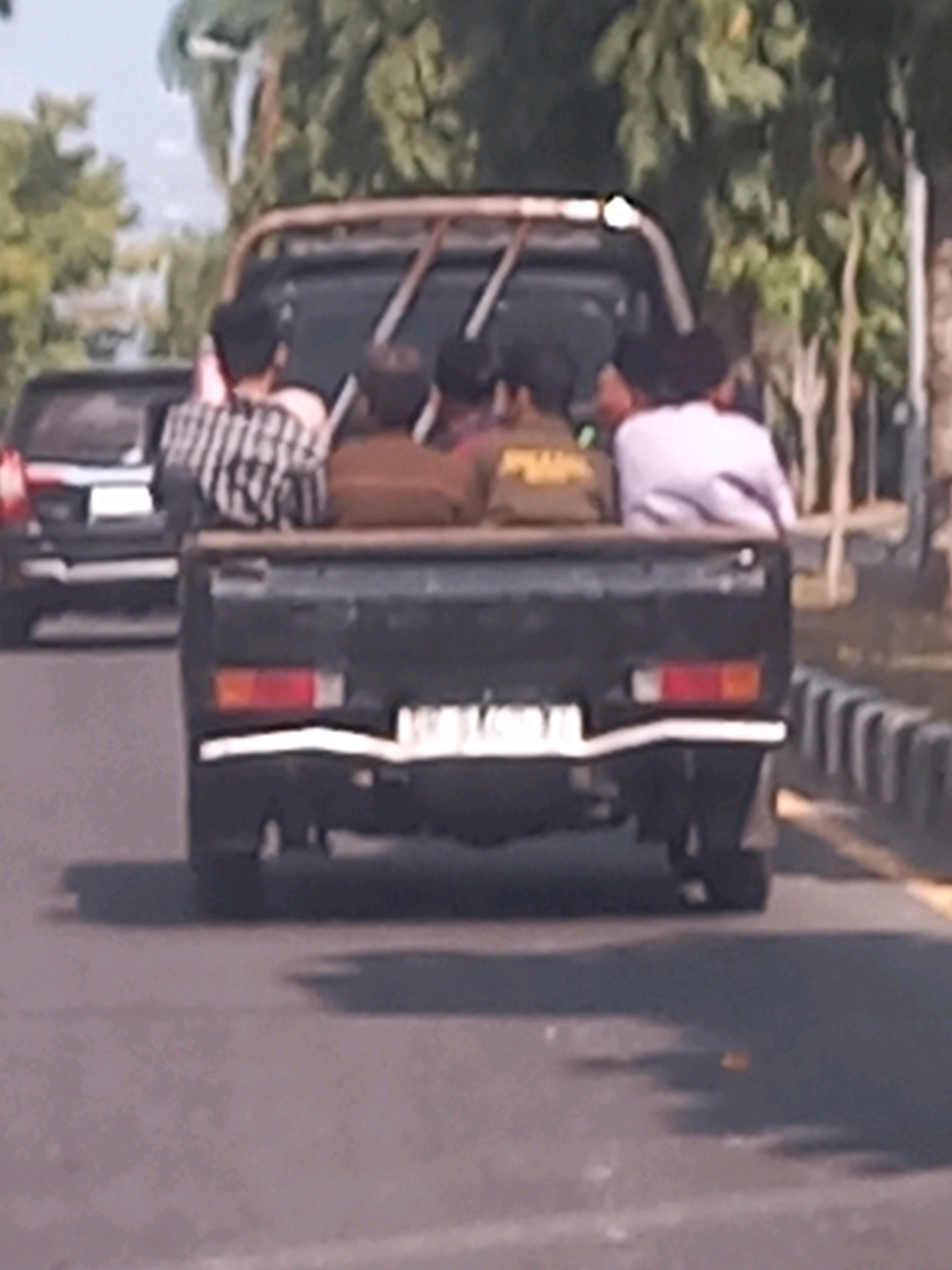 In Mataram, Indonesia, four individuals are visible in the back of a black utility pickup truck. One person in the front left of the truck bed wears a black and white striped shirt. To their right, a person in a brown shirt is seated. Behind them, two other individuals are seated, one wearing a light-colored top. The truck is on a paved road, with trees and foliage visible in the background. A white car is partially visible to the left of the truck. The scene appears to be during daylight hours.