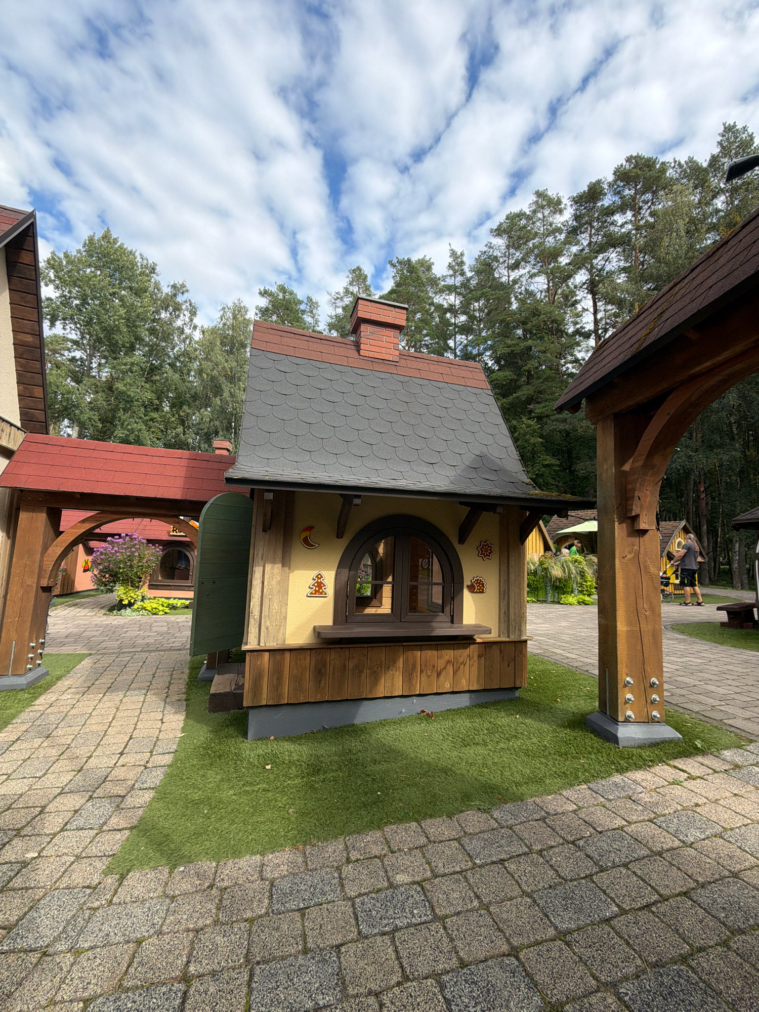 The image depicts a charming outdoor scene in the city of Tērvete, Latvia, likely within a themed park or attraction. In the foreground, a small, whimsical wooden building with a shingled, dark gray roof and a red-brick chimney stands on a patch of artificial green turf. The building has a rounded arched window with brown framing and a wooden ledge below it. The exterior walls are painted yellow with decorative motifs of a crescent moon and a gingerbread-like figure. To the left, a red-roofed structure with a wooden archway is visible, leading to more buildings in the background. The ground is paved with interlocking stone tiles, with sections of green turf interspersed. Trees with lush green foliage form a backdrop, and a cloudy blue sky with wispy white clouds is overhead. Further in the background, a man and a child are walking along the paved path, near another small, yellow building with a green umbrella. The overall atmosphere is idyllic and playful, suggesting a family-friendly environment.