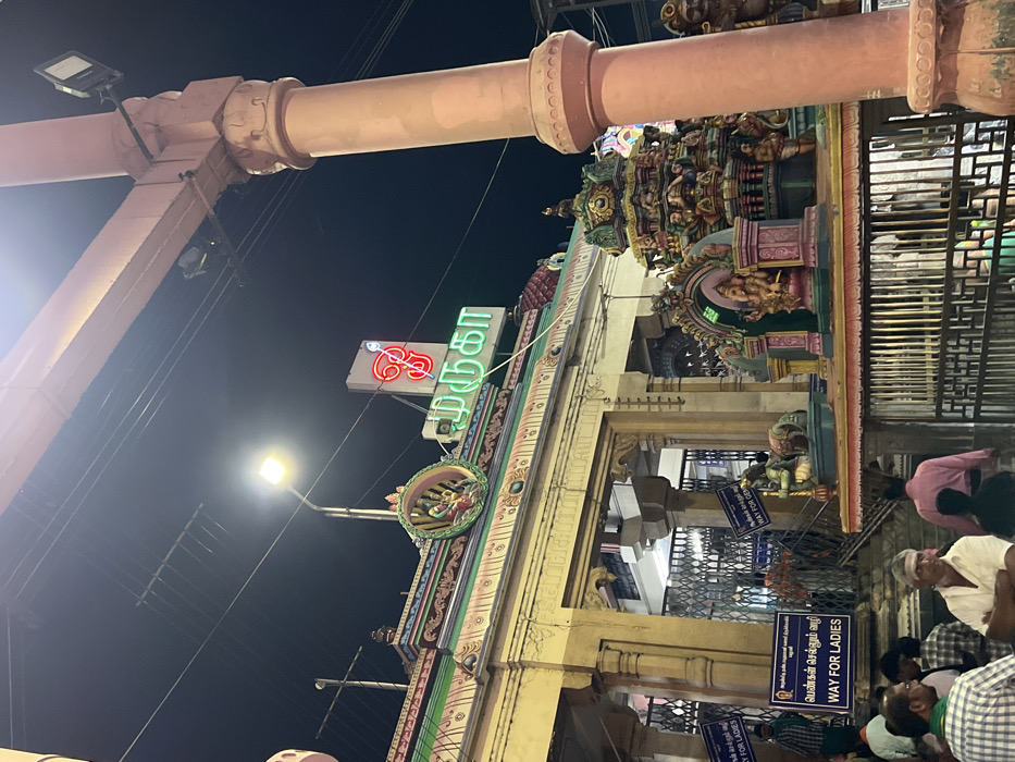 This night-time photograph captures the illuminated entrance of a Hindu temple, likely in Palani, India. The scene is dominated by a brightly lit, elaborately decorated temple gateway, adorned with colorful sculptures and architectural details. A large overhead archway or structure extends across the top of the frame.
The dark sky is punctuated by bright streetlights and prominent neon signs. A striking green and red neon sign displays the Tamil word "முருகா" (Muruga), indicating the temple's dedication to Lord Muruga, a significant deity in the region.
Numerous people are visible at the base of the temple entrance, appearing to gather or queue. Several blue and white signs are strategically placed to direct visitors. These signs prominently feature the text "WAY FOR LADIES" in English and "பெண்கள் செல்லும் வழி" (Pengal Sellum Vazhi) in Tamil, suggesting designated entry points for women. Another visible sign reads "செல்லும் வழி" (Sellum Vazhi), which means "Way" or "Path."
The atmosphere suggests an active religious gathering or a regular evening visit to the temple. The artificial lighting vividly highlights the intricate details of the temple architecture and illuminates the faces of the devotees. Metal grates or fences are also visible, possibly aiding in crowd management. The overall impression is a vibrant and spiritually significant evening at a bustling temple in Palani.