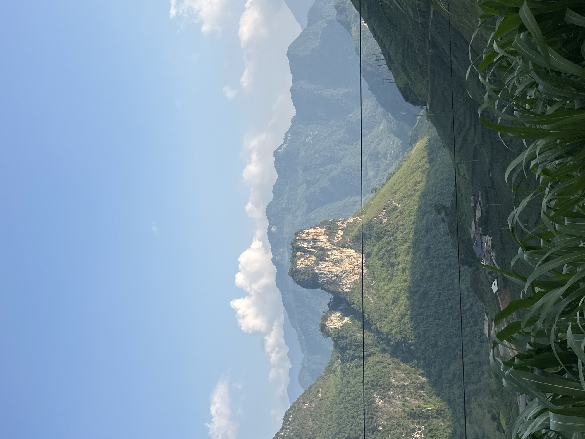 A daytime shot taken in Đường Thượng Commune, Vietnam, reveals a mountainous landscape under a blue sky with scattered clouds. A prominent rocky outcropping is surrounded by lush green vegetation on the slopes of the mountains. Power lines cut across the scene. Vegetation, possibly corn, appears in the foreground. A small village with houses is visible. Distant mountain ranges fade into a hazy background.