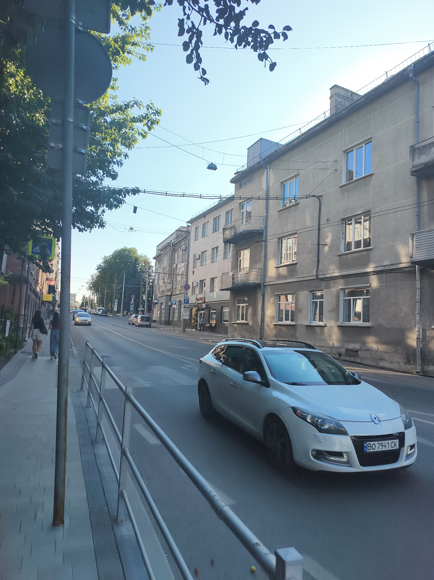A white Renault Megane is driving down a street in Ternopil, Ukraine. The car has a license plate that reads "BO 7941 CK". On the sidewalk, two women are walking away from the camera. There are buildings on both sides of the street, and trees line the sidewalk on the left. The sky is clear and blue, and the sun is shining.