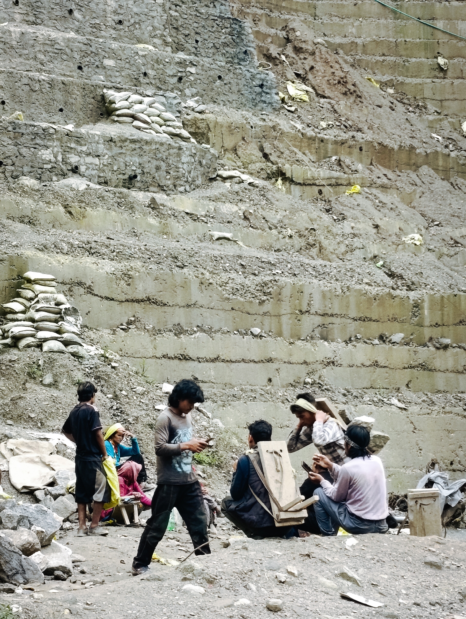 In Vashisht, India, a group of individuals is gathered at the base of a terraced, rocky embankment. Several people are seated or standing, some appearing to be engaged in conversation or resting. In the foreground, a person in a dark t-shirt and black pants is walking while looking at a mobile phone. Another individual, also in dark clothing, stands with one hand on their hip, facing away from the viewer. Further back, a person in a blue garment and yellow scarf is seated, with another person in a pink garment beside them. In the mid-ground, several individuals are interacting; one person, wearing a checkered shirt, has a wooden plank resting on their shoulders. A person in a light pink shirt and blue pants is holding a mobile phone, and another individual in a dark vest is seated next to them. The environment consists of loose rock, dirt, and a large, tiered slope constructed from concrete and soil. Sandbags are stacked in various locations along the embankment and at its base. Scattered debris, including plastic bags and what appear to be metal containers, are visible.