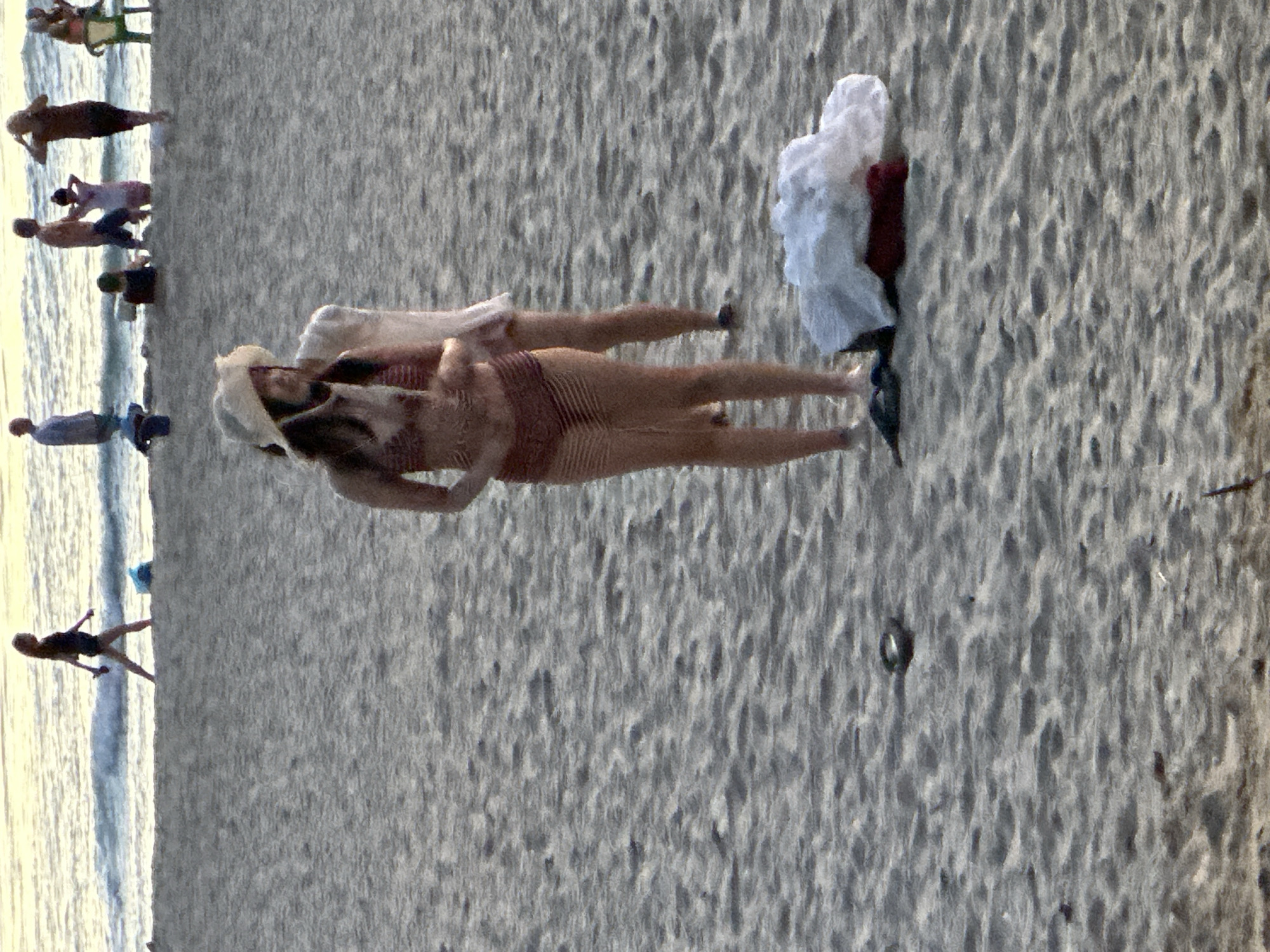 The image depicts a beach scene with several people enjoying the outdoors. In the foreground, a woman wearing a straw hat and a maroon and white striped bikini stands on the sand. Behind her, another person, partially obscured, is also on the beach. In the background, various individuals are visible near the water's edge. Some are walking along the shoreline, while others are gathered further back, possibly sitting or standing. The ocean is calm with gentle waves. The overall setting is a sandy beach with a pale sky overhead, suggesting it is either early morning or late afternoon. The specific location in Đà Nẵng, Vietnam, is not identifiable from the visual cues in the image alone.