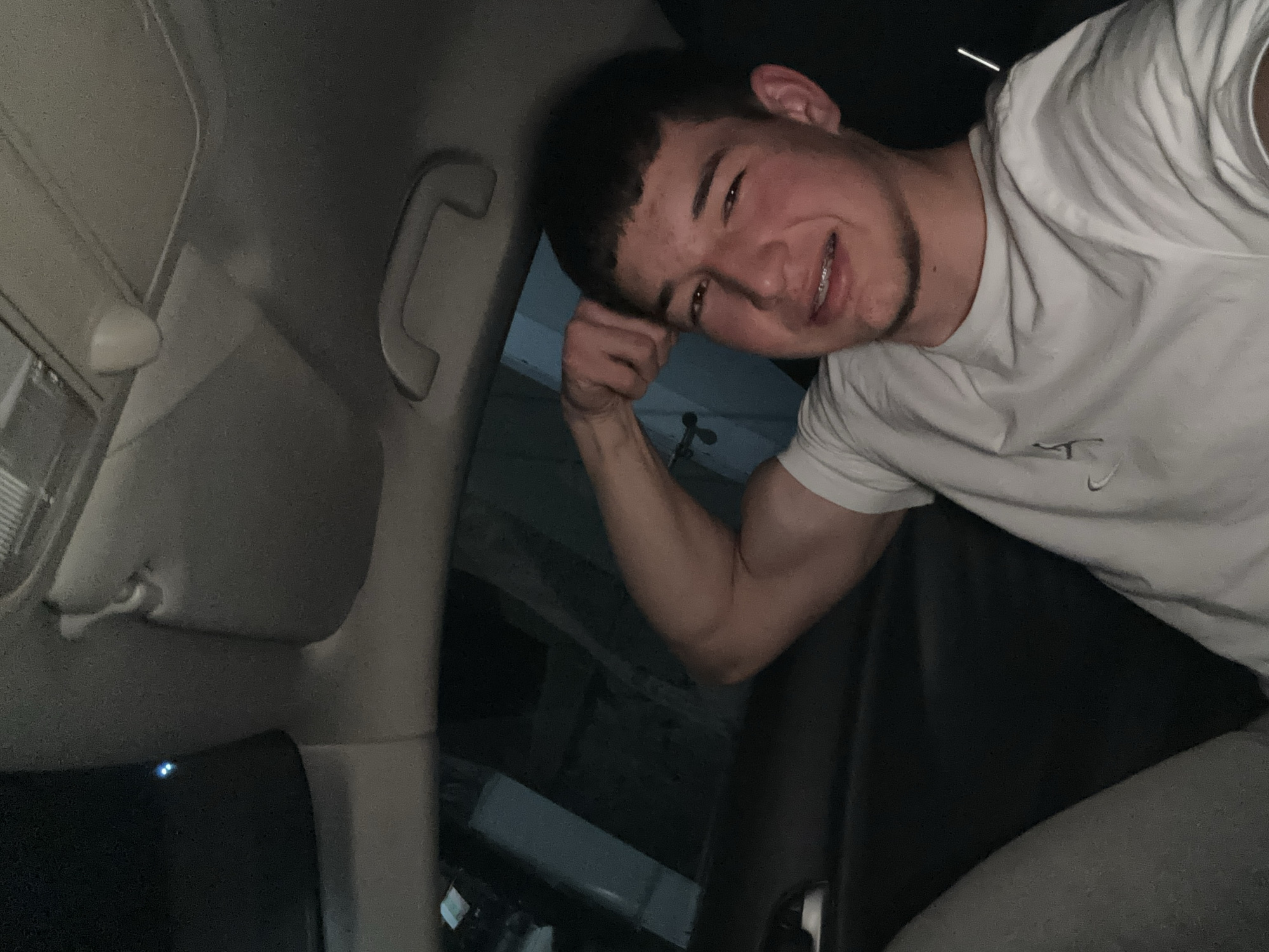 A young man is photographed inside a vehicle, possibly a car, under low lighting conditions. He is leaning his head on his left hand and smiling at the camera. He wears a white t-shirt. The interior of the car, including the headliner, sun visor, and grab handle, are visible. The specific location of the vehicle in Zlatograd, Bulgaria cannot be determined from the image.