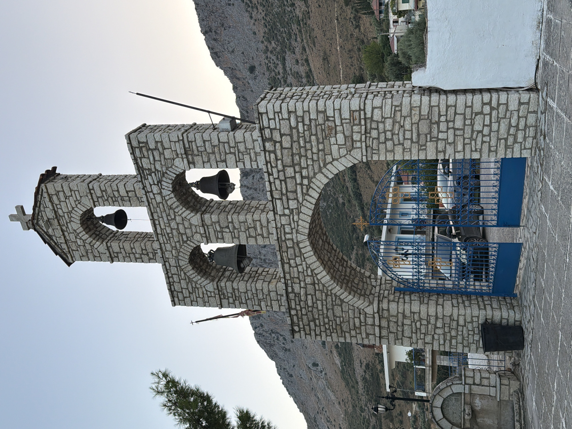 The image depicts a bell tower constructed of rough-hewn stone blocks. The tower has an arched entrance and is topped with a cross. Two large bells hang within the tower, each within an arched opening. Blue metal gates are open in front of the archway, revealing a paved courtyard and a building in the background. The bell tower is situated in a hilly, arid landscape, with a rocky mountainside visible behind it. A street lamp and a stone fountain are present to the left of the tower. The scene appears to be in the Municipal Unit of Alyzia, Greece.