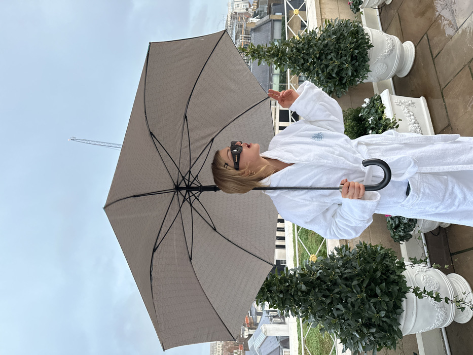 A person in a white robe holds a gray umbrella against a cloudy sky with a crane in the background. The individual wears sunglasses and makes a peace sign. The scene includes white planters with green foliage. Buildings are visible in the distance, and the setting seems to be an outdoor terrace or balcony.