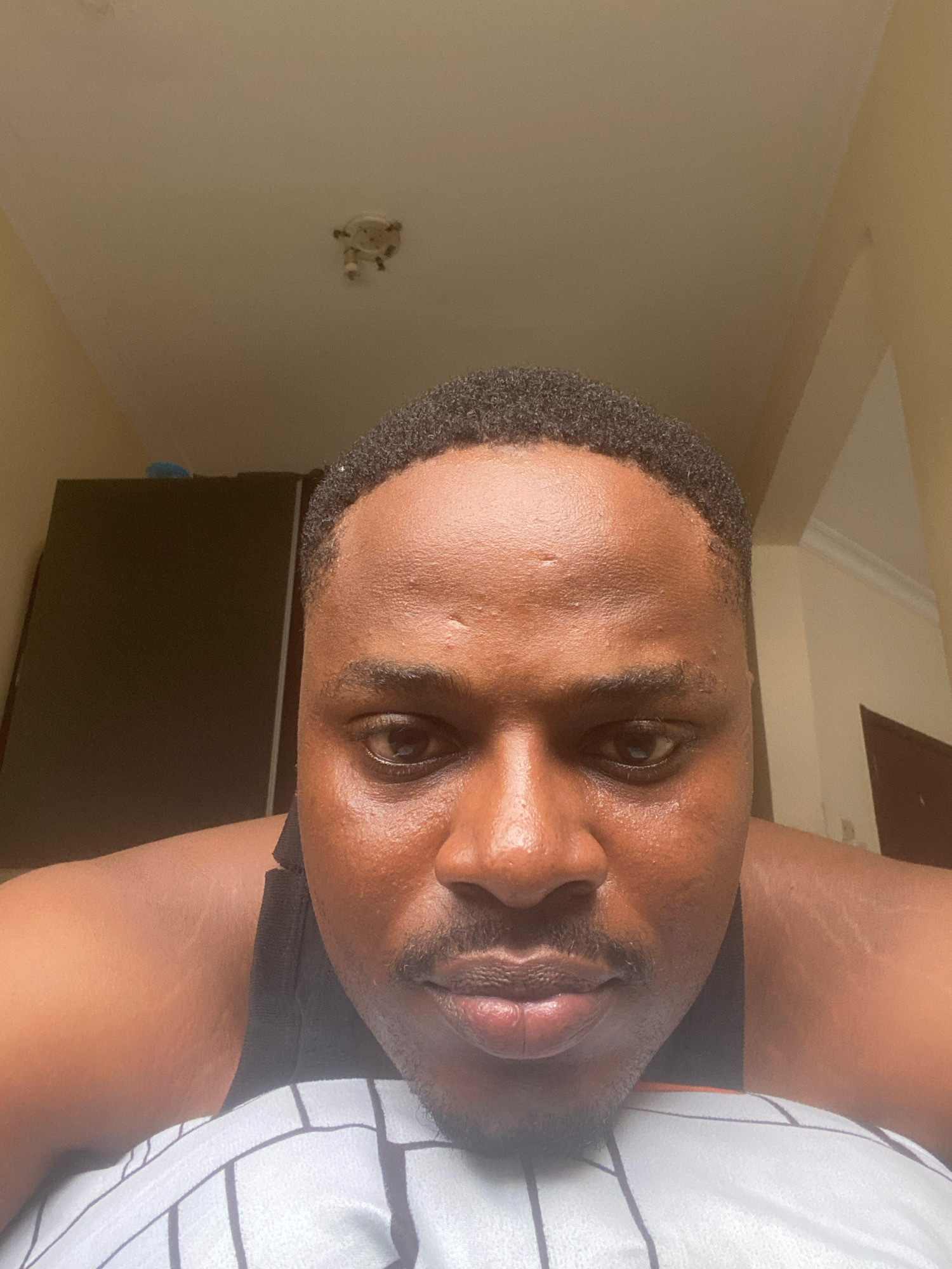 In Ojomu, Nigeria, a close-up, low-angle selfie is taken from the perspective of a man lying on his front, looking up towards the camera. The man has dark skin, short black hair, and a thin mustache and goatee. He is wearing a black tank top. The background is blurred, but appears to be an indoor setting with a ceiling light fixture visible in the upper center of the frame. To the left, a portion of a dark wardrobe is visible. The lighting suggests it is daytime, possibly with natural light entering from the side, creating some shine on the man's forehead and cheeks. The overall impression is an informal, personal snapshot.