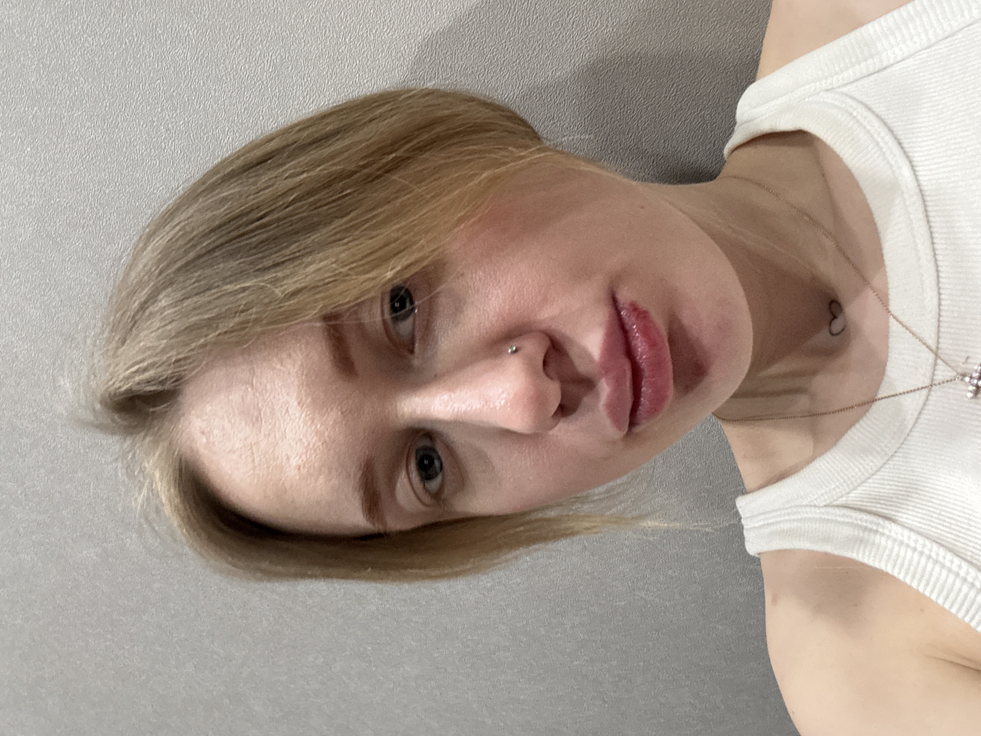 A close-up, portrait-style image, rotated 90 degrees clockwise, features a person looking directly at the viewer. The person, who appears to be female, occupies the majority of the frame. Their head is positioned towards the left side of the image, with their body extending to the right.
The person has blonde or light brown hair, styled in a way that frames their face. Their light-colored eyes (possibly blue or grey) are wide open and focused intently on the camera. A small, silver nose stud is visible on the right nostril. Their lips are full, slightly parted, and appear to have a glossy or moist finish with a reddish-pink tint. The skin has a fair complexion with visible texture, particularly on the forehead and around the eyes.
They are wearing a white, ribbed tank top or camisole, visible on the right side of the image. A delicate gold or light-colored chain necklace rests on their collarbone. A small, black outline of a heart-shaped tattoo is distinctly visible on the upper chest, slightly to the right of the center of their body.
The background is a plain, light grey surface with a subtle, fine-grained texture, possibly indicating a painted wall or wallpaper. The lighting is soft and even, highlighting the facial features without harsh shadows. The person's expression is neutral, calm, and contemplative. There is no visible text in the image.