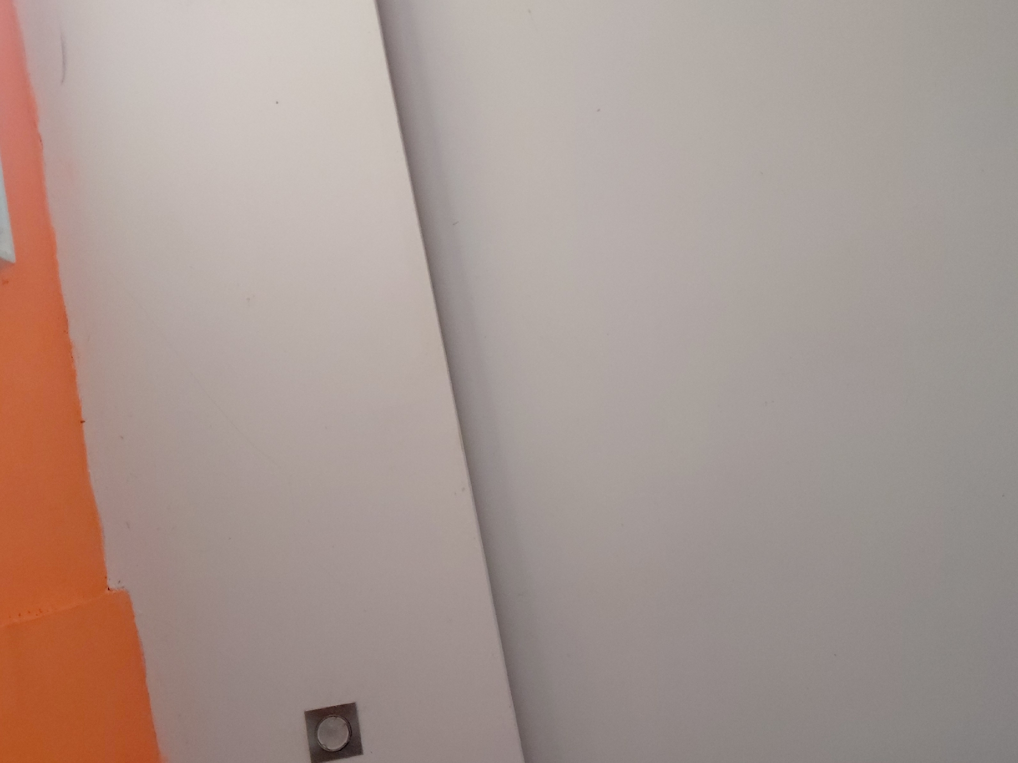 The image captures an abstract, minimalist view of an indoor architectural space in Santa Ana, Venezuela. The dominant feature is a large, smooth surface painted in a light off-white or beige color, occupying the right and central portions of the frame. A diagonal line sharply divides this main light surface from a slightly darker, possibly shadowed, section of a similar wall or structural element, running from the top-center towards the bottom-left.
On the far left, a vibrant, matte orange-reddish wall or panel extends vertically, creating a strong color contrast with the light-colored surfaces. Near the bottom-left corner of the lighter wall, a small, square-shaped fixture is visible. It appears to be made of dark metal or plastic, housing a circular, slightly convex, light-colored element in its center, resembling a flush-mounted push-button switch, a small light, or an outlet cover.
The surfaces appear plain and unadorned, with minor specks or faint marks visible on the light-colored walls. The lighting is soft and diffused, indicating an indoor setting under ambient or artificial light. No people or activities are depicted. There is no visible text. The image focuses on the interplay of geometric lines, planes, and colors within a contemporary interior design.