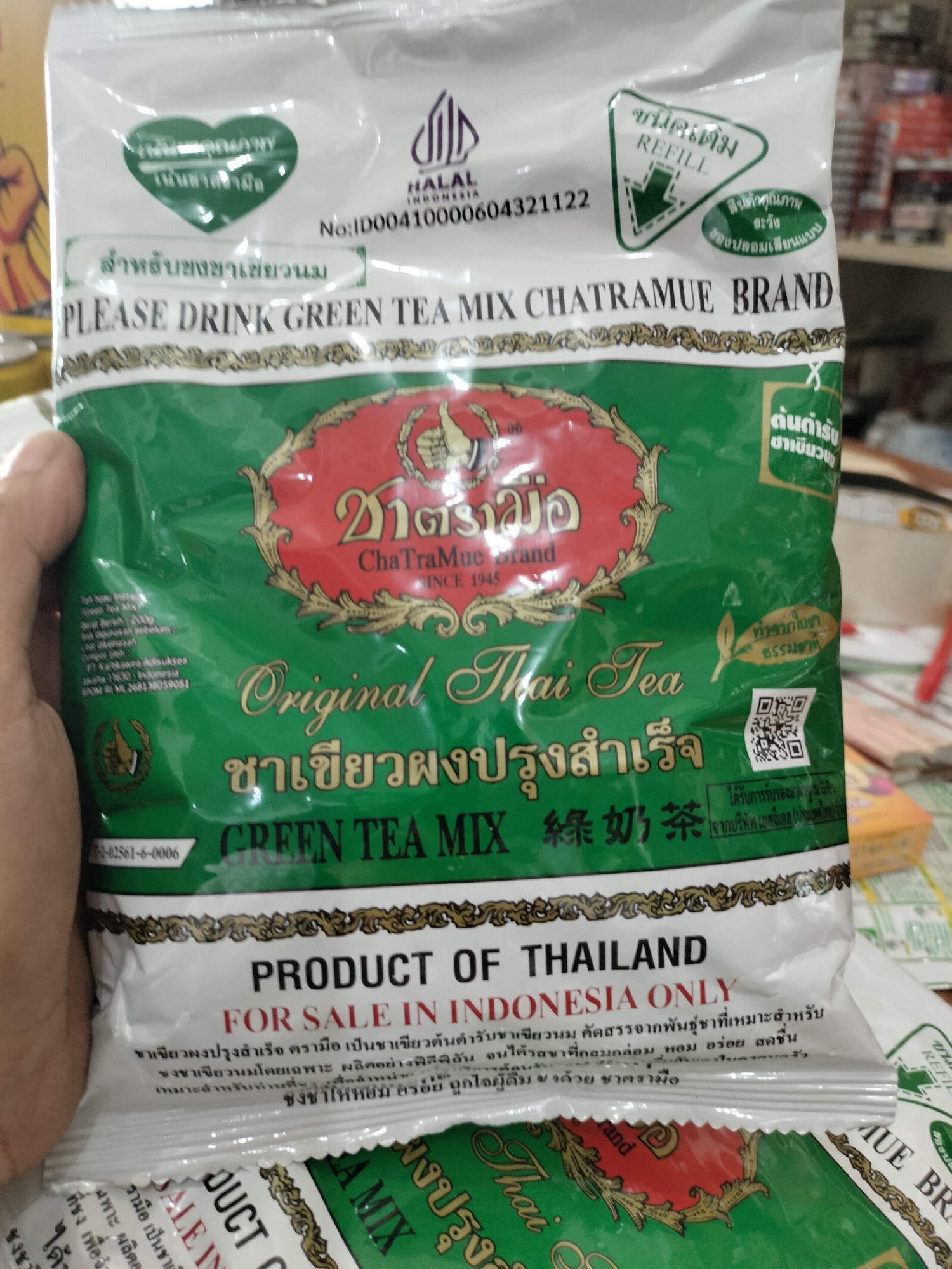 The image is a close-up, overhead shot of a green bag of "ChaTraMue Brand Original Thai Tea" mix. The bag features Thai and English text, as well as a logo and a QR code. The bag appears to be a product of Indonesia, as indicated by "FOR SALE IN INDONESIA ONLY" and "No:ID00410000604321122". The background is blurred, suggesting an indoor setting, possibly a store shelf. The image does not contain any information about Meudangara, Indonesia, or any individuals present.
