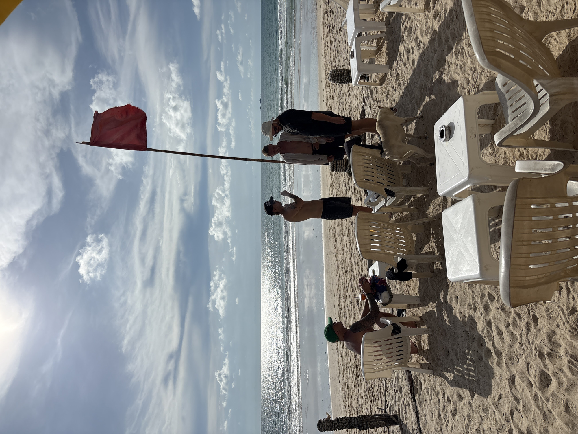 This image captures a scene on a sandy beach, likely in the city of Choeng Thale, Thailand, on a day with a partly cloudy sky and bright sunshine reflecting off the ocean. In the foreground, several white plastic chairs and small white tables are arranged on the sand, suggesting an outdoor seating area for a beachside establishment.
In the mid-ground, four individuals are present. On the left, a person wearing a green cap is seated in a chair, holding a cigarette and what appears to be a drink. To their right, three men are standing and conversing. One man, wearing a black cap, is gesturing with his hand. Another man is shirtless, and the third is wearing a light-colored shirt and a hat. A light-colored dog is standing near the group of men.
A prominent red flag is flying on a pole behind the standing men, indicating potential ocean conditions. The ocean stretches out to the horizon, with gentle waves lapping the shore. The beach itself is sandy, with footprints and textures visible. The overall atmosphere suggests a relaxed day at the beach.