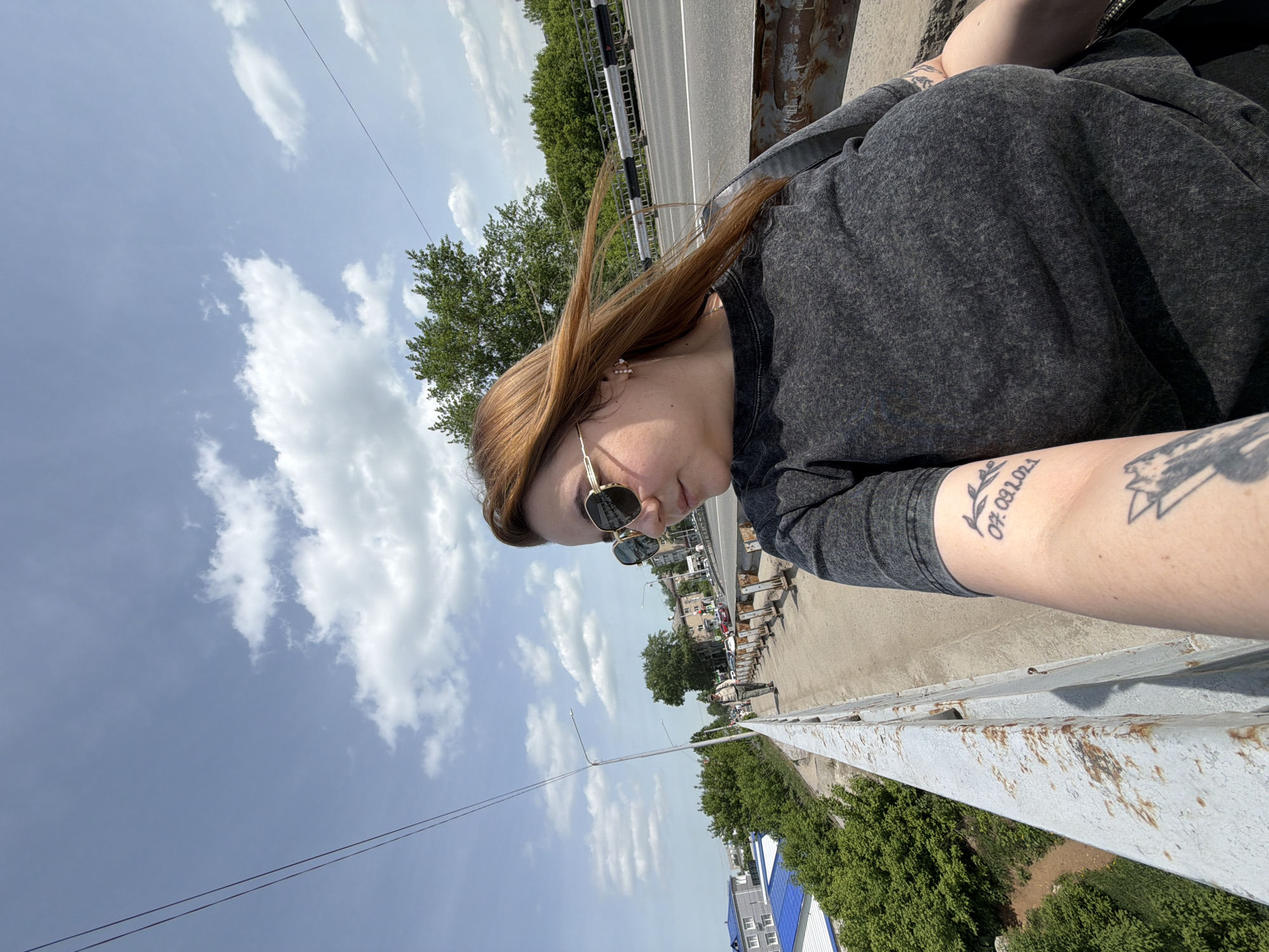 A woman with fair skin, auburn hair, and sunglasses is captured in a medium, off-center shot. She wears a gray sweatshirt and has tattoos on her right forearm. Her left arm rests on a gray metal bridge railing.
Behind her and to the left is a daytime sky with scattered white clouds. The scene overlooks the city of Chaikovsky, Russia. Trees, buildings, and a road with traffic are visible in the background. The viewpoint is likely from the bridge over the Saygatka River, a bridge connecting Chaikovsky to the village of Saygatka.