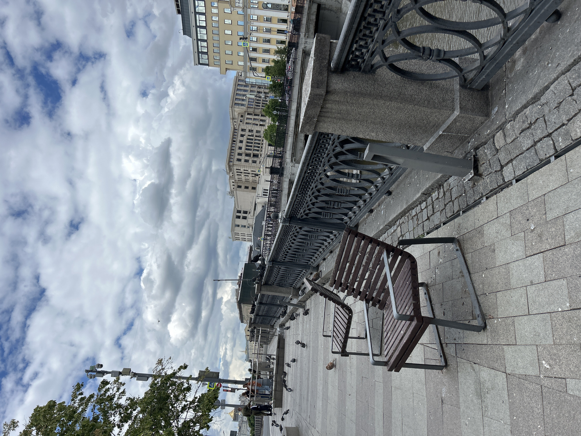 The photograph shows a section of a city embankment, likely in Moscow, Russia, with overcast skies. A row of dark-colored, ornate metal fencing runs along the edge of the embankment, with grey stone support posts spaced at intervals. Adjacent to the fencing, there are several wooden benches with metal frames positioned on a paved walkway.
Below the embankment, there is a lower level where people can be seen walking, standing, and interacting, seemingly feeding pigeons. There is a streetlight pole with traffic signals to the left of the frame. In the background, there are several multi-story buildings with light-colored facades, typical of urban architecture. The ground is paved with stone tiles.