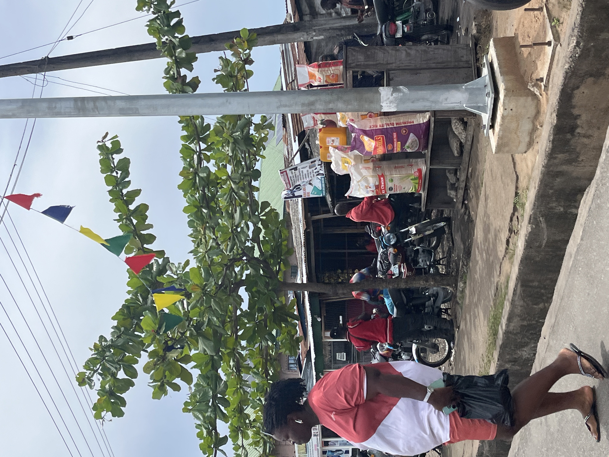 The image shows a street scene in Badagry, Nigeria. A woman in a red and white shirt, black shorts, and sandals is walking along the sidewalk. She is holding a phone in her hand. Next to her, there is a parked motorcycle. Overhead, there are trees decorated with colorful triangular flags, as well as power lines and a light pole. Behind the motorcycle, there is a small shop with bags of rice and other items displayed. Other parked motorcycles are also visible. The environment appears to be a bustling urban street.