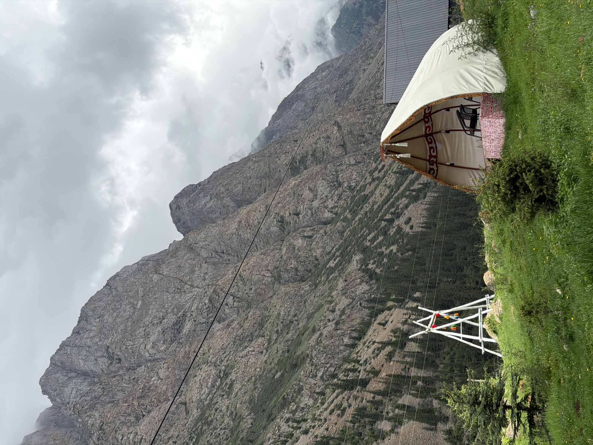 This image depicts a yurt situated on a grassy hillside with a dramatic mountain backdrop. The yurt, a traditional nomadic dwelling, is partially open, revealing a chair and a patterned rug inside. The exterior of the yurt is a light color, possibly canvas, with decorative trim. The surrounding landscape is characterized by steep, rocky mountains shrouded in mist and clouds, suggesting a cool or overcast day. The green grass on the hillside is lush, with scattered small yellow flowers. A white, A-frame structure, possibly a power line support, is visible in the foreground, adorned with colorful elements. The overall scene evokes a sense of remote, natural beauty, and a sense of peacefulness or solitude. The lighting suggests it is daytime, but the cloudy conditions create a diffused light. The rocky texture of the mountains and the presence of the yurt are strong indicators of a mountainous, possibly high-altitude, region. There is no visible text in the image.