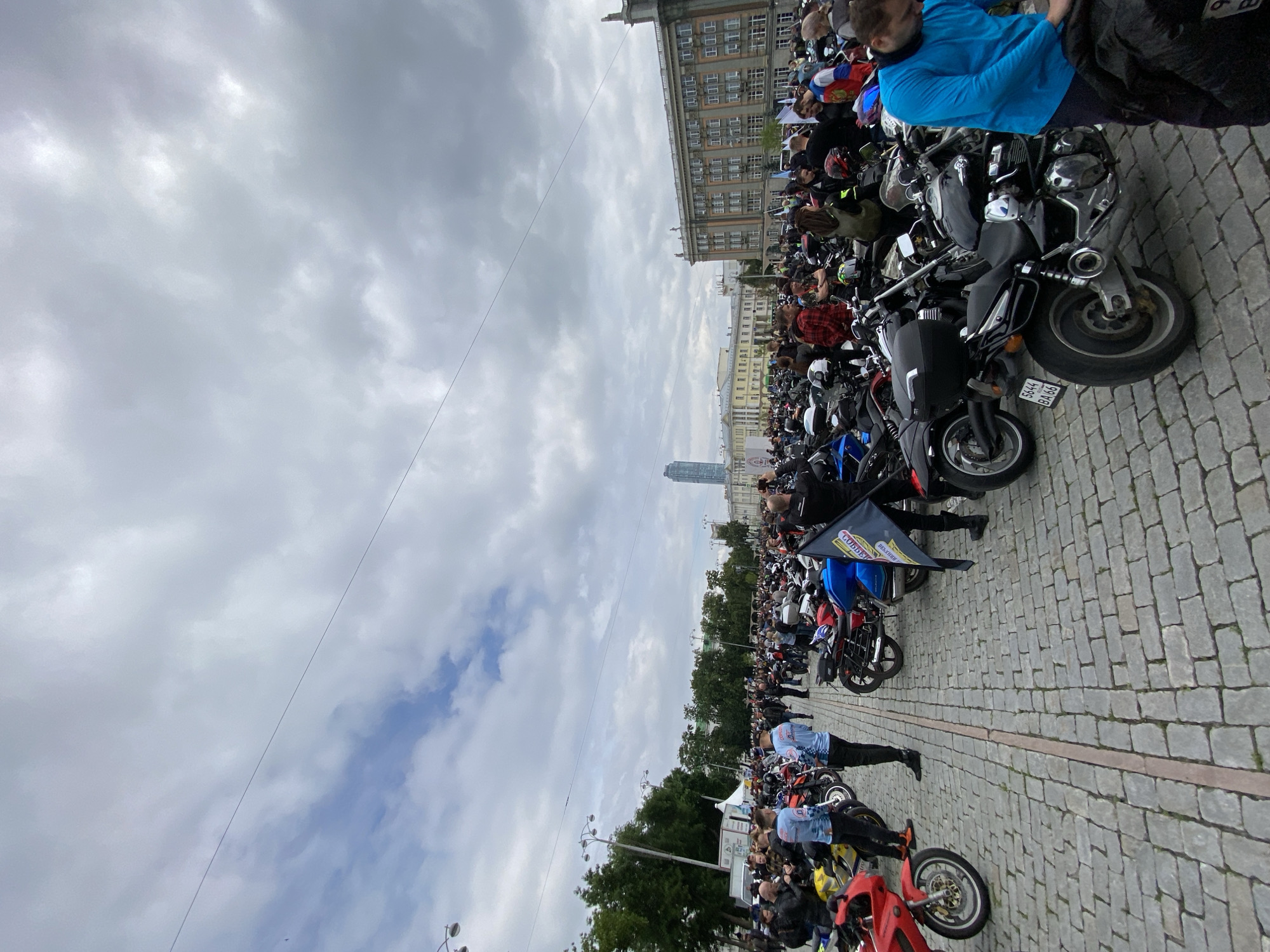 This image captures a large outdoor gathering of numerous individuals and motorcycles on a cobblestone-paved square under an overcast sky. The scene is located in Yekaterinburg, Russia, identifiable by the prominent Vysotsky Business Center skyscraper visible in the distant background, left of center, and historical architectural buildings, one notably with rooftop statues, on the right. A license plate "5644 BA 66" on a black motorcycle further confirms the Sverdlovsk Oblast region (code 66).
In the immediate foreground, grey cobblestone pavement with a distinct central dark line extends into the midground. A red motorcycle's front wheel and part of its engine are visible on the far left. On the right, a black cruiser-style motorcycle with a rear top case and the aforementioned Russian license plate is parked prominently. A man in a bright blue long-sleeved shirt is attending to a black bag near this motorcycle.
The midground is densely populated with a crowd of people and various motorcycles, predominantly cruisers and touring bikes, parked closely together. Individuals, mainly adults, are standing, conversing, and observing the motorcycles. Many wear casual clothing, while some are dressed in motorcycle gear, including jackets and helmets. A person near the center holds a dark flag with yellow lettering or a logo. Another person on the right wears a jacket with white, blue, and red segments.
In the background, beyond the immediate gathering, rows of green trees are visible on the left. Several multi-story buildings of varying architectural styles line the horizon. To the left, a large, modern glass skyscraper (Vysotsky Business Center) stands tall. To the right, two large, classical-style administrative or university buildings feature numerous windows, ornate cornices, and statuary on the roofline of the building on the far right. The sky is entirely cloudy, presenting a uniform grey tone with some brighter patches, indicating diffused daylight. No direct sunlight is visible. Overhead, thin wires traverse the sky.