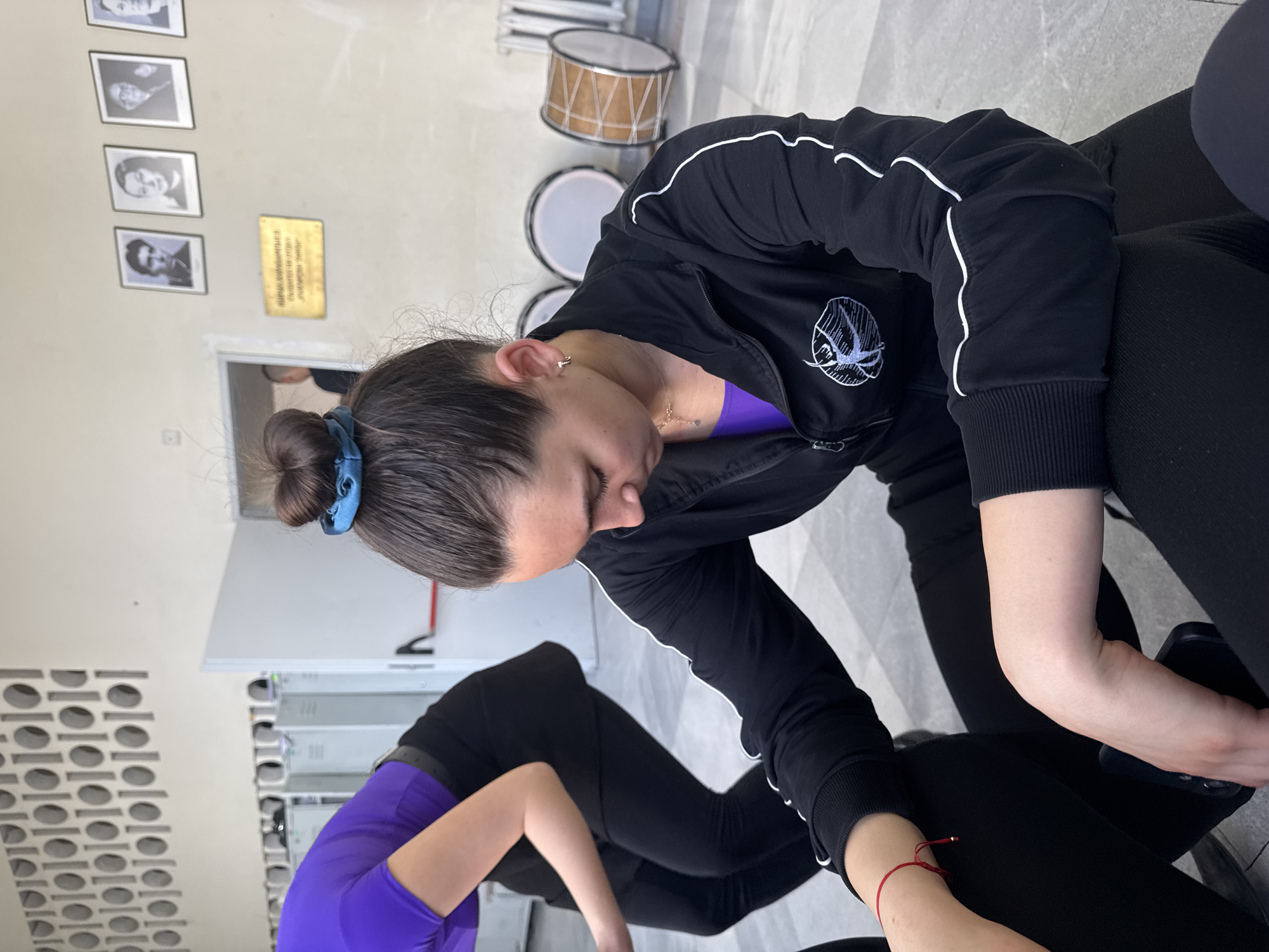 This media file captures an indoor scene, likely a practice or studio space in Sofia, Bulgaria. The central subject is a person with dark hair tied in a bun with a blue scrunchie, positioned in the foreground and looking downwards. They are wearing a black zip-up athletic jacket with white piping, a purple top underneath, and black leggings. A small hoop earring and a delicate necklace are visible, and a thin red string bracelet adorns their left wrist. The jacket features a stylized white logo, resembling a basketball or a round object with radiating lines, on the left chest.
Partially visible in the bottom left is another person, also dressed in a purple top and black leggings. In the background, against a light-colored wall, four framed black and white portraits are arranged vertically, depicting individuals who appear to be historical or cultural figures. Adjacent to these portraits, a golden plaque with multiple lines of text written in Cyrillic script is affixed to the wall.
Further back in the room, several drums are visible, one with a wooden finish and others with white heads, suggesting a musical or performance context. A patterned wall section and some metallic shelving or lockers are also present. The floor is a light, possibly tiled surface. The scene conveys a sense of quiet focus, possibly during a break or preparation for an activity. The lighting suggests an indoor setting during the day, likely with artificial illumination.