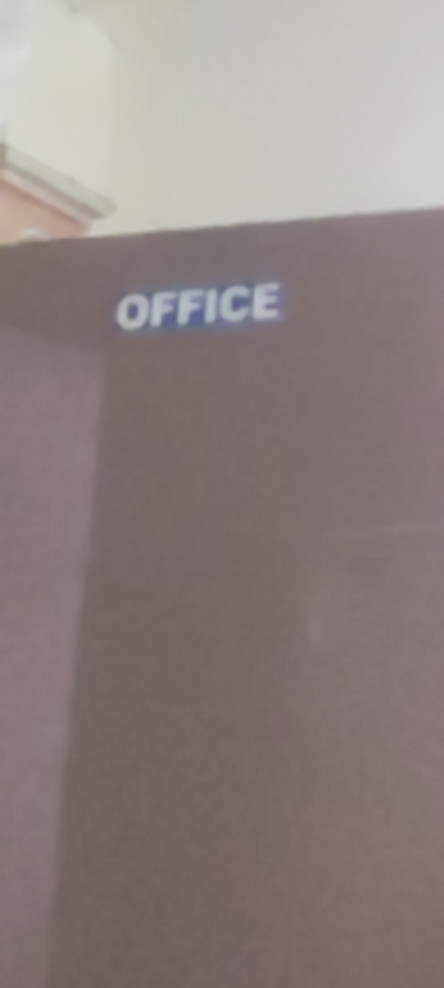 The image shows a close-up, slightly blurry view of a dark brown surface with the word "OFFICE" printed in blue and white lettering. The lettering is capitalized and appears to be embossed or applied as a decal. The word is positioned in the upper left quadrant of the visible surface. The background above the surface is out of focus and appears to be a light-colored, possibly white or off-white, ceiling or wall. The scene is static and does not contain any people, actions, or interactions. There is no information in the image to suggest a location in Gebang, Indonesia.