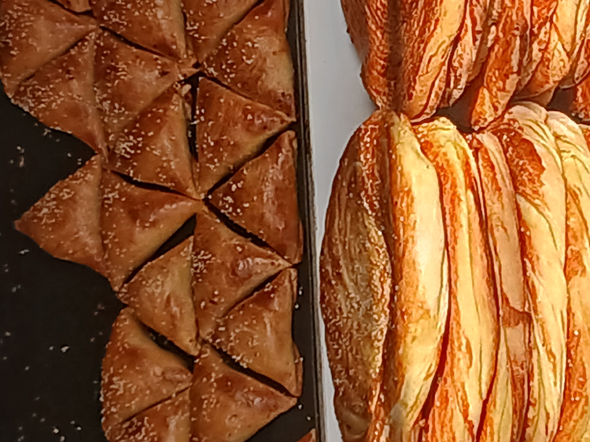 The image displays a close-up view of baked goods, likely pastries, arranged in two distinct tiers. The upper tier features numerous triangular-shaped pastries, each sprinkled with what appears to be sesame seeds. These pastries are golden brown and arranged in a dense, tiled pattern. Below this, the lower tier showcases larger, layered pastries with a flaky appearance, also golden brown and sprinkled with seeds. The overall impression is of a bakery display or a collection of freshly baked items. The image does not provide any contextual information regarding its specific location in Khankiz, Uzbekistan.