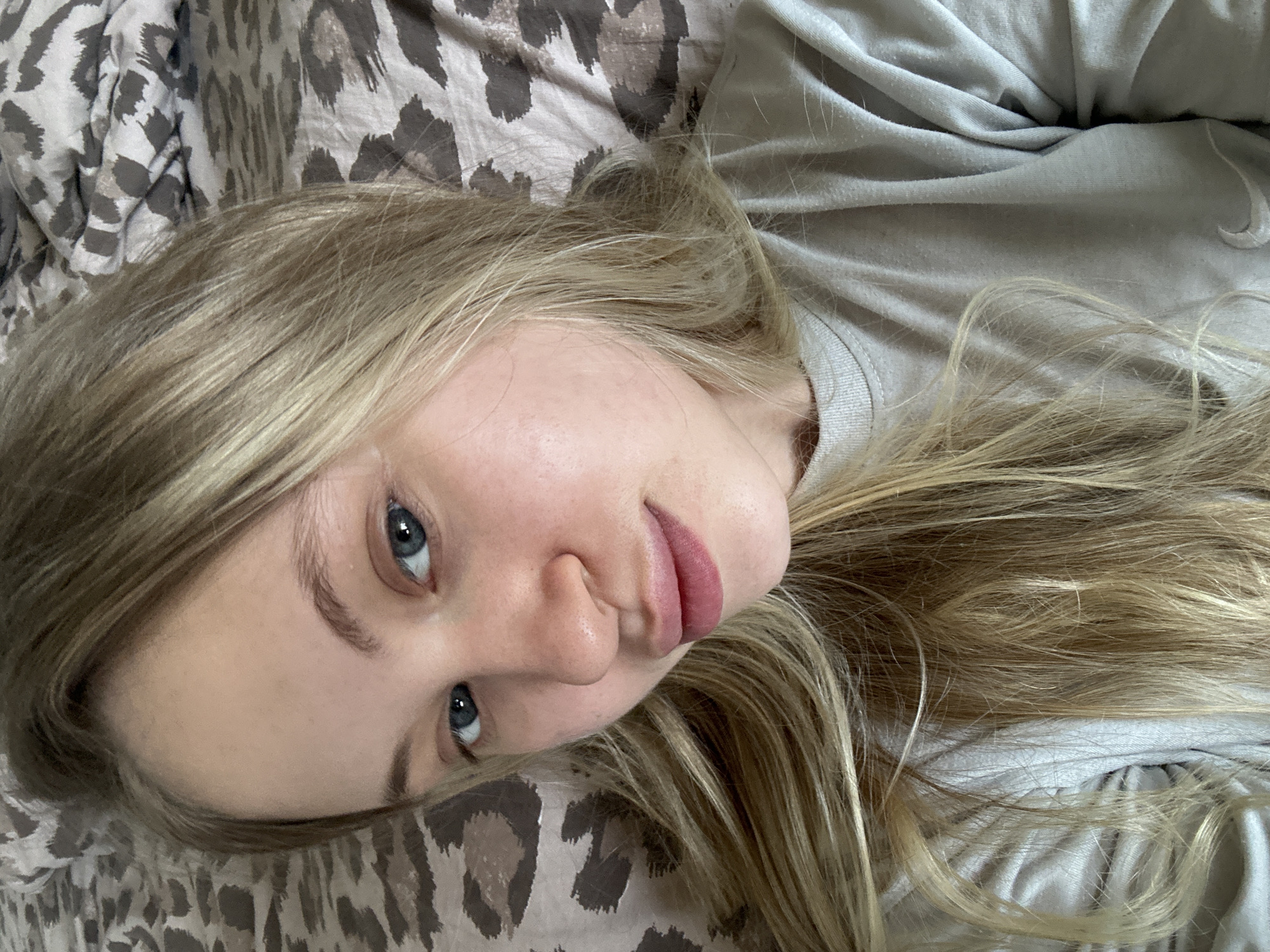 A young woman with blonde hair and blue eyes lies on a bed with leopard-print bedding. She is wearing a light-colored t-shirt. The image is a close-up selfie. The scene appears to be indoors, likely a bedroom. The image is located in Miass, Russia.