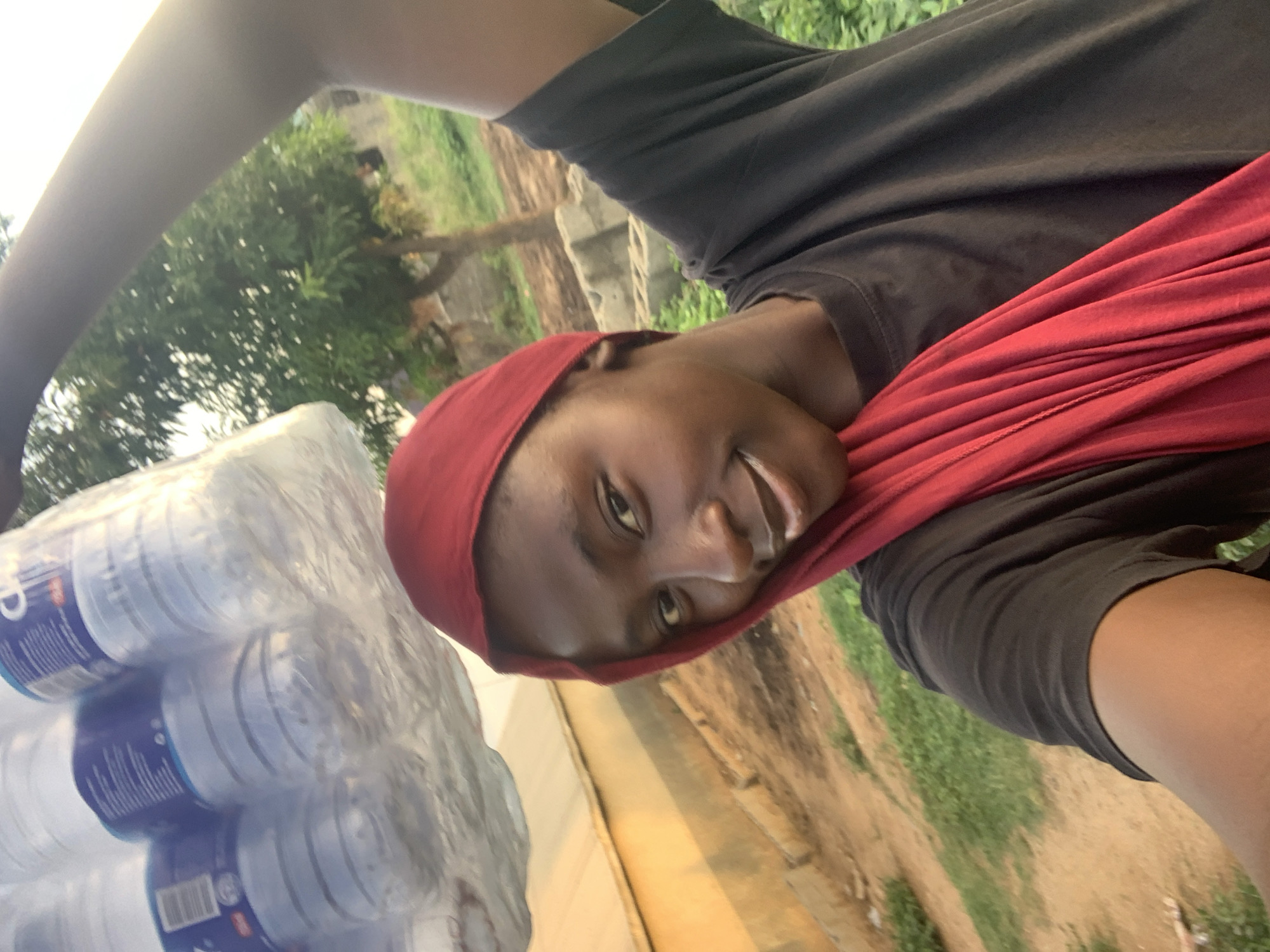 A person is holding a large stack of water bottles, presumably in the city of Keffi, Nigeria, based on the user request. The individual, wearing a red headscarf and a dark shirt, appears to be taking a selfie or posing for a photo. A red cloth hangs around their neck. The background shows trees, vegetation, and what seems to be a wall or a fence. The ground appears to be dirt or grassy.