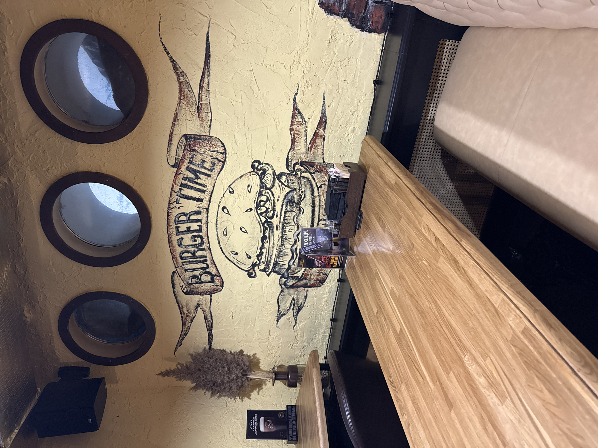 This image depicts the interior of what appears to be a restaurant or bar, likely located in Russia given the visible text. The main subject is a textured, light yellow wall decorated with a large, stylized mural. The mural features a banner displaying the English text "BURGER TIME" alongside an illustration of a hamburger with clock hands on its top bun, suggesting a temporal theme for the eatery. Abstract, flame-like designs in dark brown and red surround the burger and banner.
To the left of the mural, three dark-framed circular windows, resembling portholes, are embedded in the wall. Their reflections indicate they look into an adjacent interior space rather than outside. A black speaker or electronic device is mounted on the wall below the lowest porthole.
Running along the right side of the image is a light-colored wooden table, suggesting a dining area. Behind the table, a plush, light-colored tufted sofa or bench seating provides comfort. On the table, various items are visible: a holder containing menus or promotional cards, a stack of black napkins, and a small vase with dried, feathery decorative plants.
The visible text on the promotional materials on the table includes "Новый BEER HOL" (New Beer Hall), "ЗАХОДИ, пи что...?" (Come in, drink what...?), and on a black card, "ДОЛЖЕН БЫТЬ Stout" (Must be Stout) with a graphic of a stout beer glass. Below this, a standard Russian warning reads: "ЧРЕЗМЕРНОЕ УПОТРЕБЛЕНИЕ АЛКОГОЛЯ ВРЕДИТ ВАШЕМУ ЗДОРОВЬЮ" (Excessive consumption of alcohol harms your health).
The scene implies a casual dining or drinking atmosphere, possibly a themed burger and beer establishment. The lighting is artificial, providing no indication of the time of day or outdoor conditions. No individuals are present in the frame.