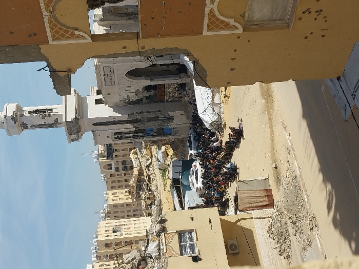 The image depicts a heavily damaged urban environment in Khan Yunis, Palestinian Territory, under a clear blue sky. In the immediate foreground on the right, a multi-story, light-colored building shows significant structural damage, with a large section of its upper wall completely missing, exposing internal rebar. Multiple circular impact marks, consistent with bullet or shrapnel damage, are visible on the remaining facade.
In the mid-ground, approximately 100-150 individuals, predominantly men, are seated or kneeling in organized rows on unpaved, dusty ground. They appear to be facing forward. Located directly behind and to the left of this gathering are several white tents, including one larger dome-shaped structure, and two white SUVs. Behind the assembled individuals, a prominent mosque with a tall, white minaret is visible. Both the mosque's main structure and its minaret show extensive damage, with large sections of material missing, exposing underlying construction, and numerous impact points. Arabic script is discernible above the mosque's arched entrance.
The background is filled with numerous multi-story residential buildings, primarily light tan or yellow, many of which exhibit severe damage, including collapsed sections, missing walls, and visible rubble. Piles of debris and construction rubble are scattered across the ground throughout the scene, particularly near the damaged buildings and along an unpaved road in the lower mid-ground.
