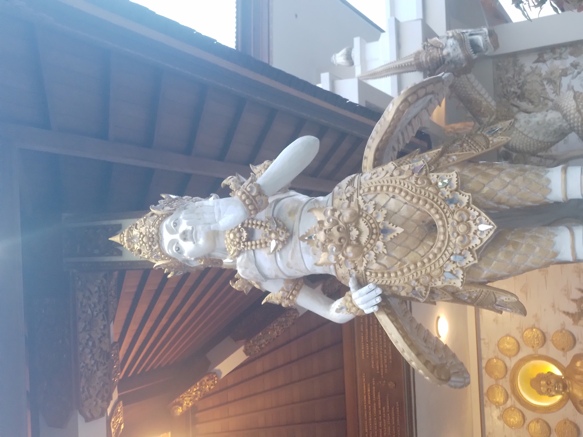 The image shows an ornate, likely religious, statue in Kuta Selatan, Indonesia. The statue is predominantly white and gold, depicting a figure with elaborate headwear and detailed clothing. Part of a building is visible in the background, featuring a wooden ceiling and what appears to be a plaque with text. There is also a display of circular objects, possibly coins or medallions, arranged around a central figure. The scene is well-lit, although the image is rotated, making the composition slightly awkward.