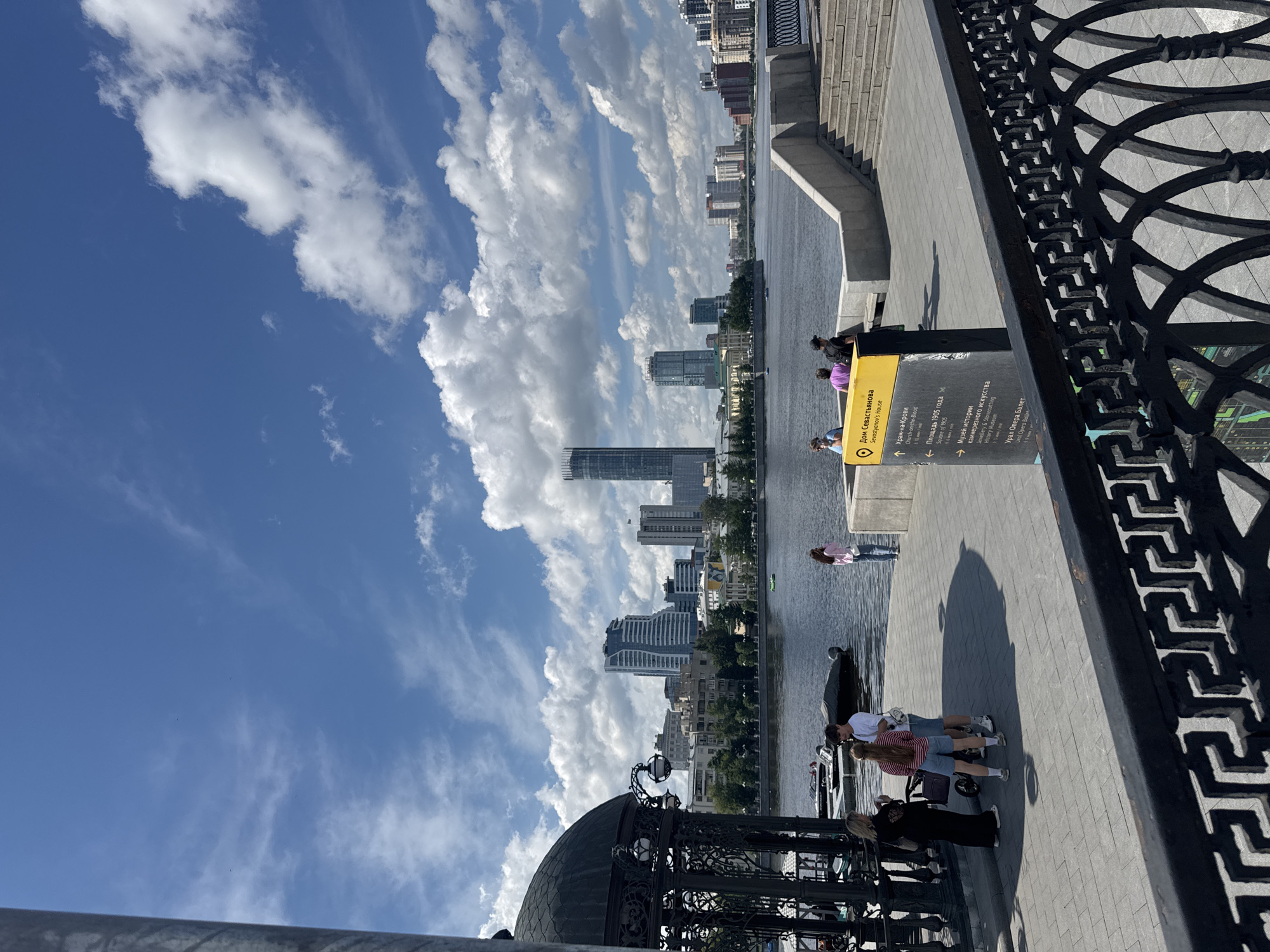 This is a daytime, eye-level shot taken from a waterfront promenade in Yekaterinburg, Russia, likely along the Iset River. In the foreground, a black ornamental metal railing with a geometric pattern separates the viewer from a paved walkway. To the left, a dark, ornate gazebo with a domed roof and decorative lampposts is partially visible. Several people are present on the promenade: a woman in a black dress and headscarf, a couple with a stroller, and a woman in a pink shirt and blue jeans standing near the water. A yellow information sign with Russian text, including "Дом Севастьянова" (Sevastyanov's House), is positioned on the promenade.
In the middle ground, a wide river flows under a bright blue sky dotted with white, fluffy clouds. Several boats, including a small green one and a covered motorboat, are on the water. The opposite bank of the river is lined with a cityscape featuring various buildings, trees, and a pedestrian walkway with stairs leading up from the water.
The background is dominated by a panoramic view of Yekaterinburg's modern skyline, characterized by numerous high-rise buildings of varying architectural styles, including a prominent tall skyscraper with a slender, almost needle-like top. The overall scene depicts a lively urban waterfront on a clear, sunny day.