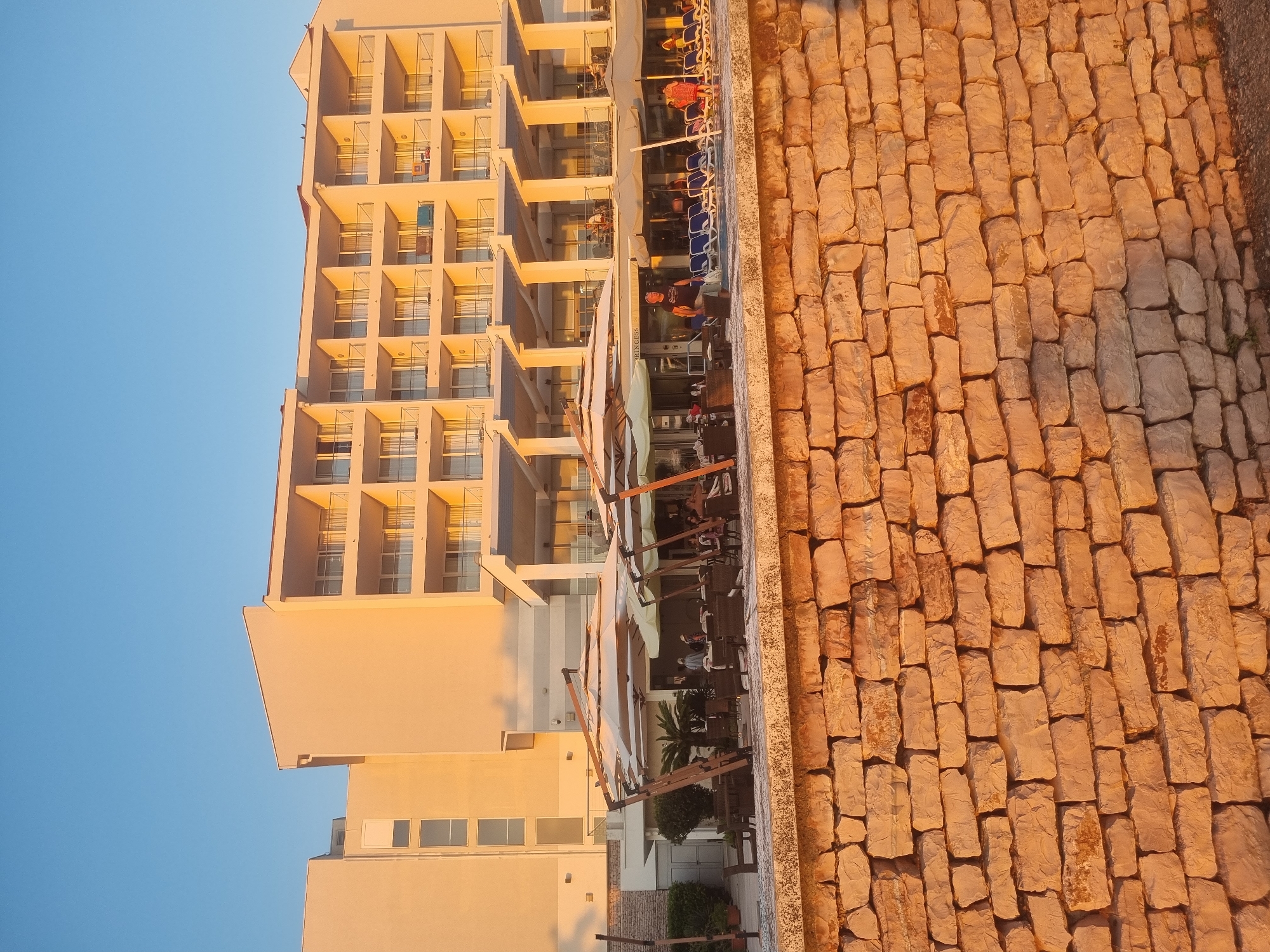 The image depicts a waterfront hotel or resort in Bar, Montenegro, bathed in the warm, golden light of either sunrise or sunset. A substantial stone wall, constructed from rough-hewn, rectangular blocks, dominates the foreground and extends across the lower portion of the frame. Behind the wall, a paved area is visible, with several tables and chairs set out under large, angled umbrellas, suggesting an outdoor dining or seating area. Beyond this, a swimming pool with blue lounge chairs is partially visible. The hotel itself is a multi-story building with a light-colored facade and numerous balconies, many of which are equipped with railings. Some people are visible on the balconies and near the pool area, indicating activity within the resort. The sky above is a clear, vibrant blue. The overall impression is one of a serene coastal resort environment.