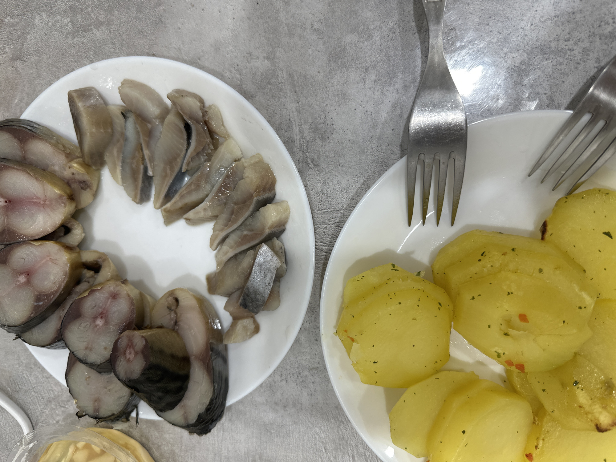 The image shows two white plates on a gray surface. The plate on the left contains slices of smoked mackerel and herring. The plate on the right contains boiled potatoes with herbs. Two forks are near the plate of potatoes. The location is identified as Chaikovsky, Russia, but no explicit details in the image confirm the location.