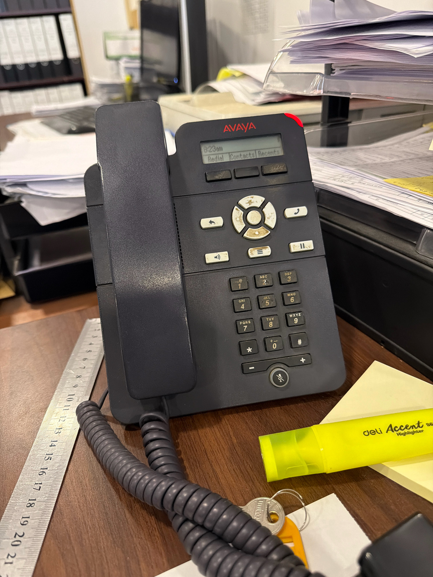 The image shows a desk with a dark gray Avaya desk phone prominently displayed in the foreground. The phone has a small screen displaying "8:23am" and options like "Redial," "Contacts," and "Recents." Below the screen is a keypad with numbers and symbols, and several function buttons. A coiled phone cord extends from the base of the phone. To the left of the phone, a metal ruler with inch and centimeter markings is visible. In the background, stacks of papers and binders are present, suggesting an office environment. A yellow highlighter pen and a set of keys are also on the desk. The lighting suggests an indoor setting, possibly an office in Riffa, Bahrain, given the reference to the city. There are no people visible in the image.
