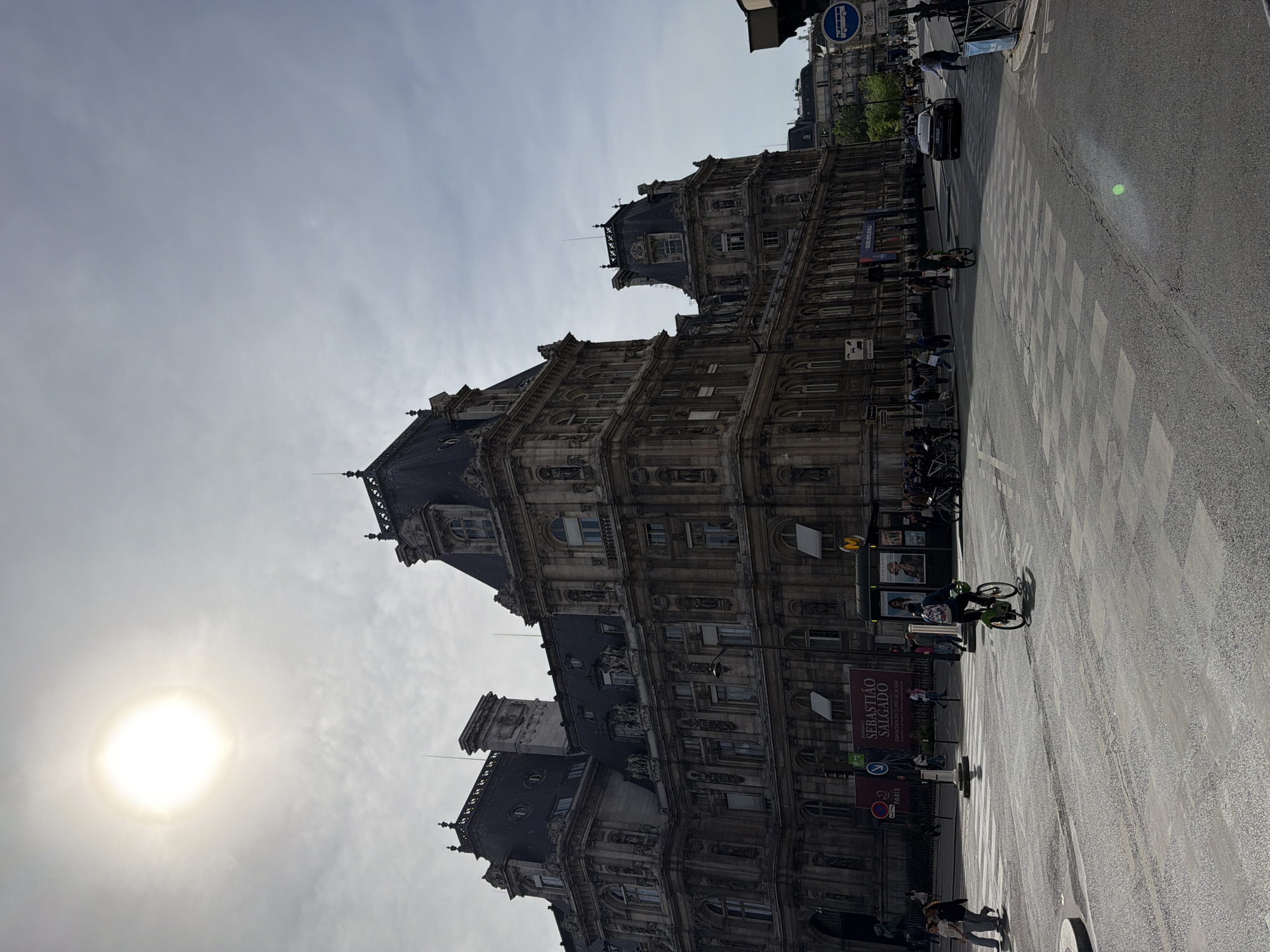 The image captures a grand, ornate historic building under an overcast sky, with a bright sunburst visible on the left side. The multi-story building, characteristic of Parisian architecture, features numerous windows, intricate sculptural details, and dark slate roofs topped with decorative finials.
Adjacent to the building is a busy urban street scene. An asphalt road with a prominent zebra crossing extends across the foreground and midground. Several people are visible, including pedestrians walking on the sidewalk and at least two cyclists on the street. A dark car is also present, moving on the road.
Notable details include the bright, almost halo-like sun obscured by clouds, indicating daytime conditions. Various signs provide context: a circular blue sign with white bus and bicycle symbols (indicating a bus lane or route), a red "P" sign with "PARIS" below it, and a yellow "M" symbol (likely for the Métro). Most prominently, a large poster on the building advertises an exhibition for "SEBASTIÃO SALGADO" with the French text "EXPOSITION | L'ESTHETIQUE D'UN REGARD" (EXHIBITION | THE AESTHETIC OF A GAZE). The scene depicts a lively street in a European city, consistent with Paris, France.