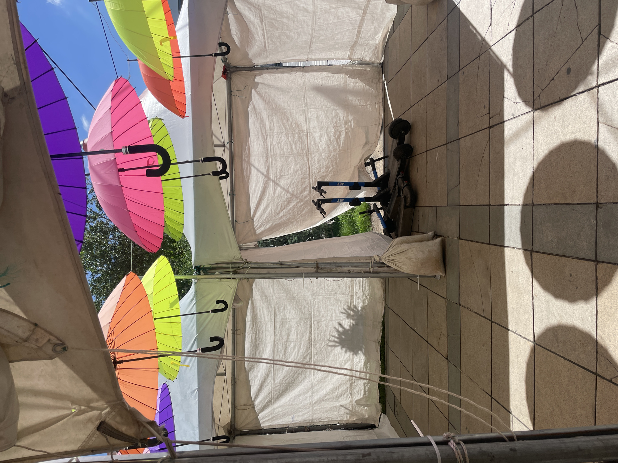 This image depicts an outdoor scene under a series of white tents, possibly at a temporary market or event. Colorful umbrellas are suspended from the tent ceilings, including purple, orange, lime green, and pink. Two electric scooters, one blue and one gray, are parked inside one of the tents on a tiled floor. A sandbag rests against a tent pole, and there are ropes and shadows visible. The scene appears to be located in the city of Ulaanbaatar, Mongolia.