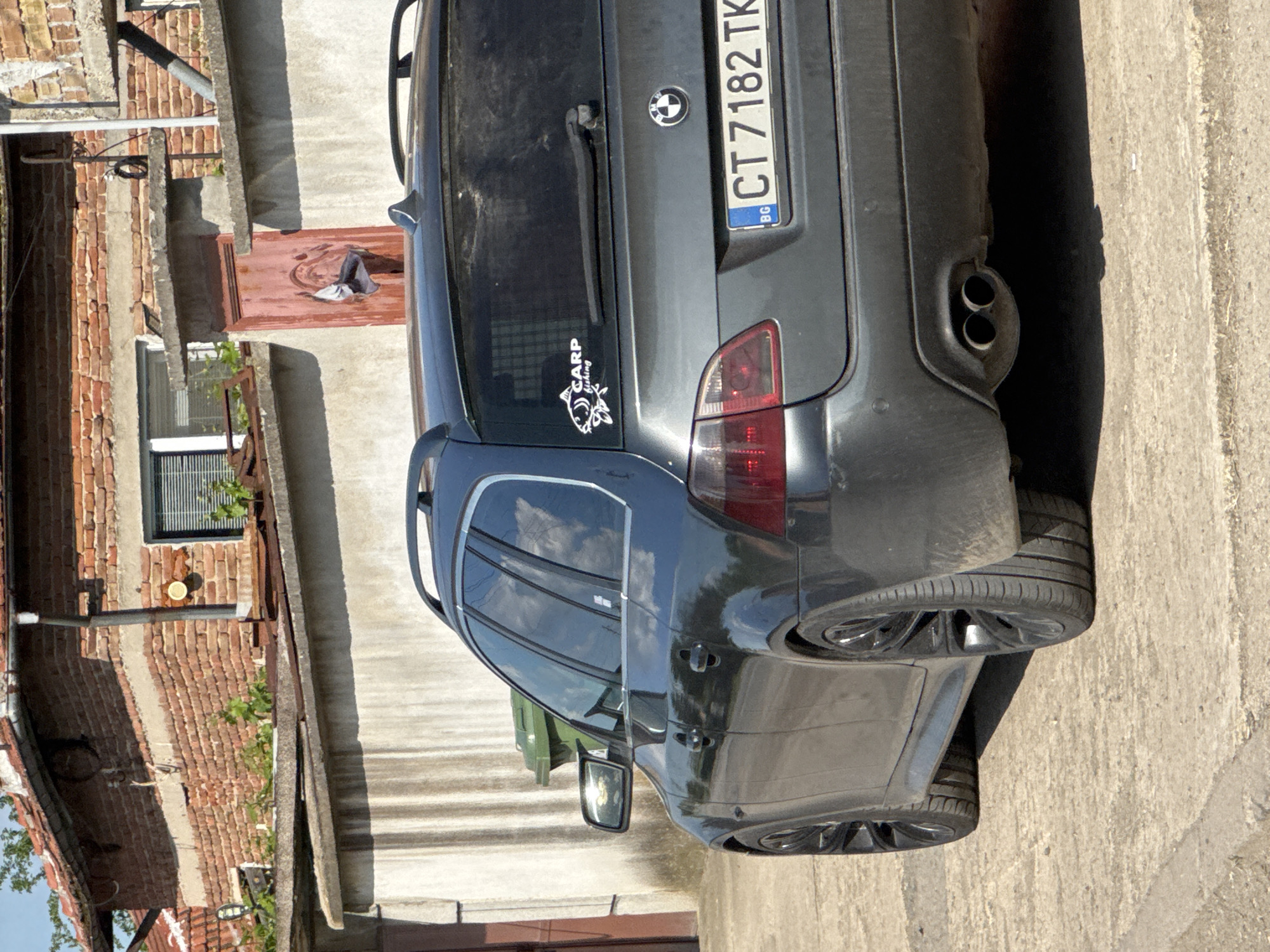 The image shows the rear of a dark grey BMW station wagon, parked outdoors, possibly on a residential street. The car has a license plate reading "CT 7182 TK" and a "CARP fishing" sticker on the rear window. The car appears to be in Sarnevo, Bulgaria, indicated by the "BG" on the license plate. The background includes a building with a brick facade, a window, and what appears to be a partially visible open doorway. The environment is a combination of urban and residential elements. The presence of stairs suggests varying terrain levels. There are no people visible in the immediate scene.