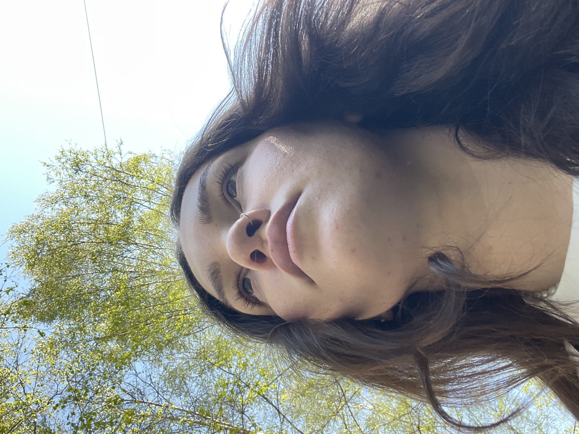 The image is a low-angle selfie of a young woman with light skin, brown hair, and blue eyes. She has a nose ring in her left nostril. The background features a clear, light blue sky and green tree branches with leaves. There is a slight blur, particularly in the background. The focus is on the woman's face. The lighting appears to be natural daylight. There is no specific information in the image to indicate that it was taken in Izhevsk, Russia. Therefore, it's not possible to ascertain the location based on visual details alone.