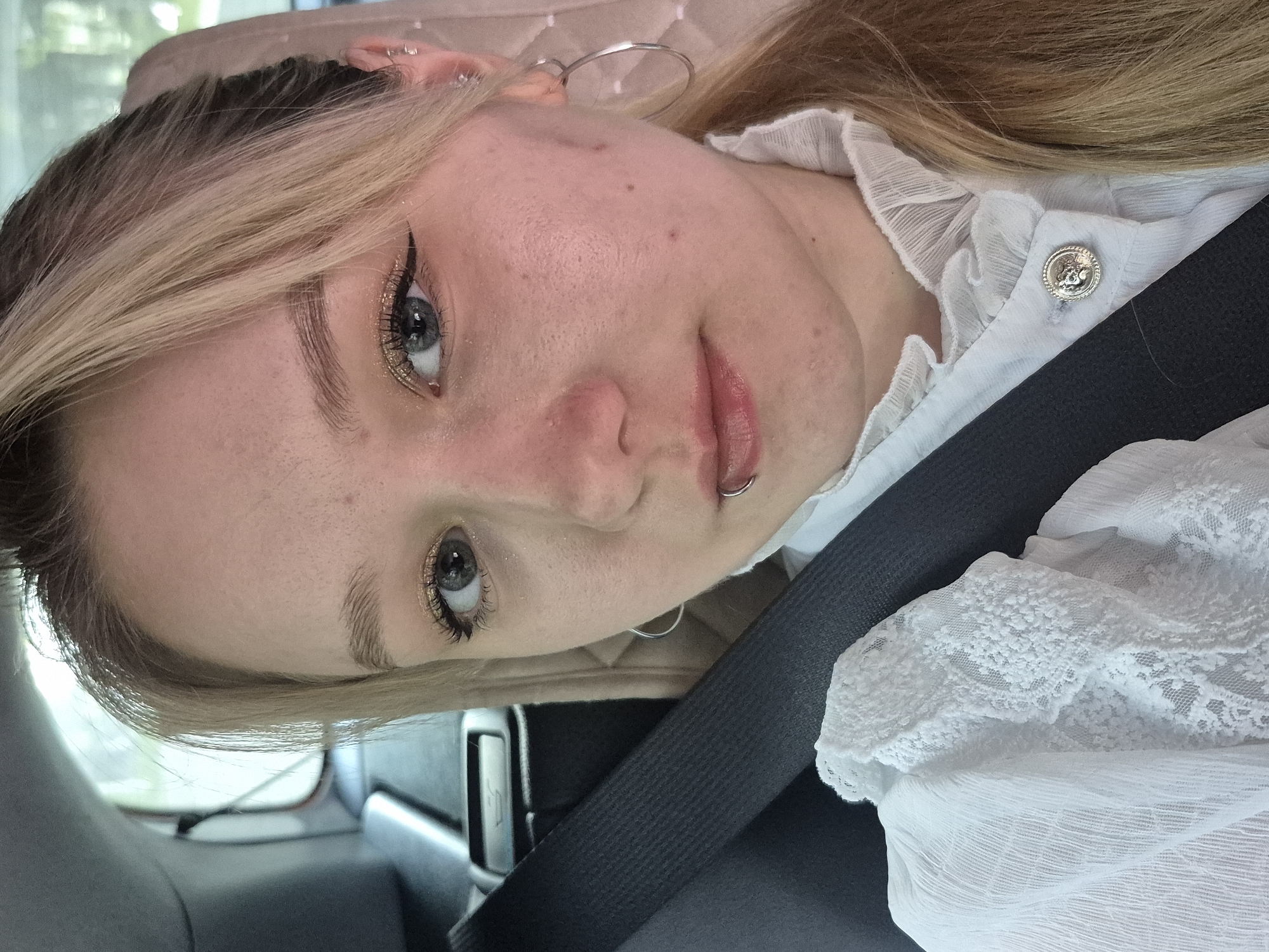 A young woman with blonde hair, wearing a white ruffled blouse, is in the passenger seat of a car, looking at the camera. She has on makeup, including eyeliner and glitter eyeshadow, and multiple ear piercings and a lip ring. A seatbelt is visible across her chest. The background shows a car interior and a blurred view of greenery outside the window, suggesting a daytime setting. The image is cropped to focus on the woman's face and upper body. No specific details of Chaikovsky, Russia are visible.