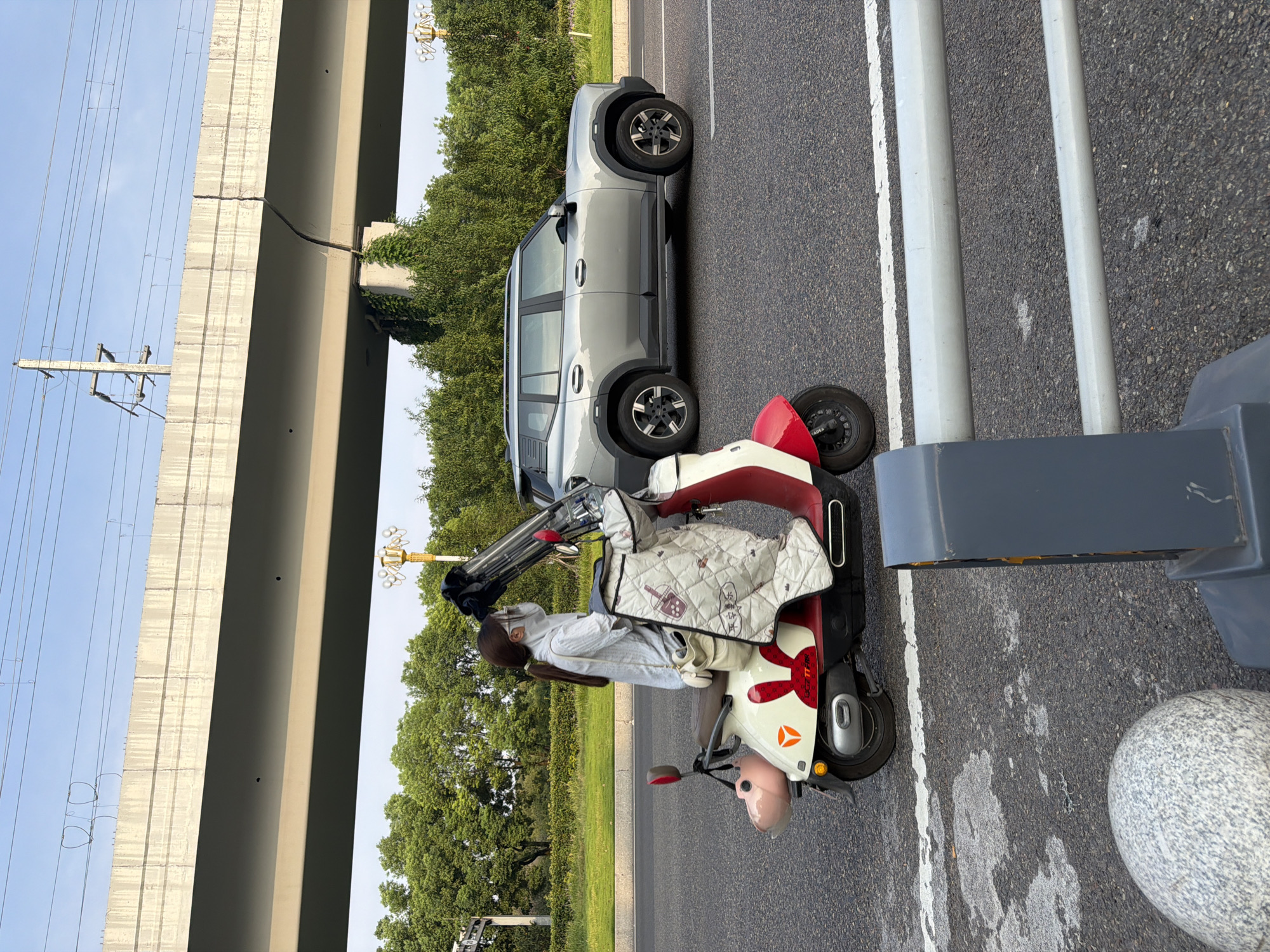 A woman wearing a face mask and a clear face shield rides an electric scooter along a road in Jiangjin, China. The scooter is red and white with a decorative blanket covering its front. A light-colored SUV drives behind her. In the background, a concrete overpass supports electrical lines, and lush green trees line the sides of the road. The sky is a clear blue.
