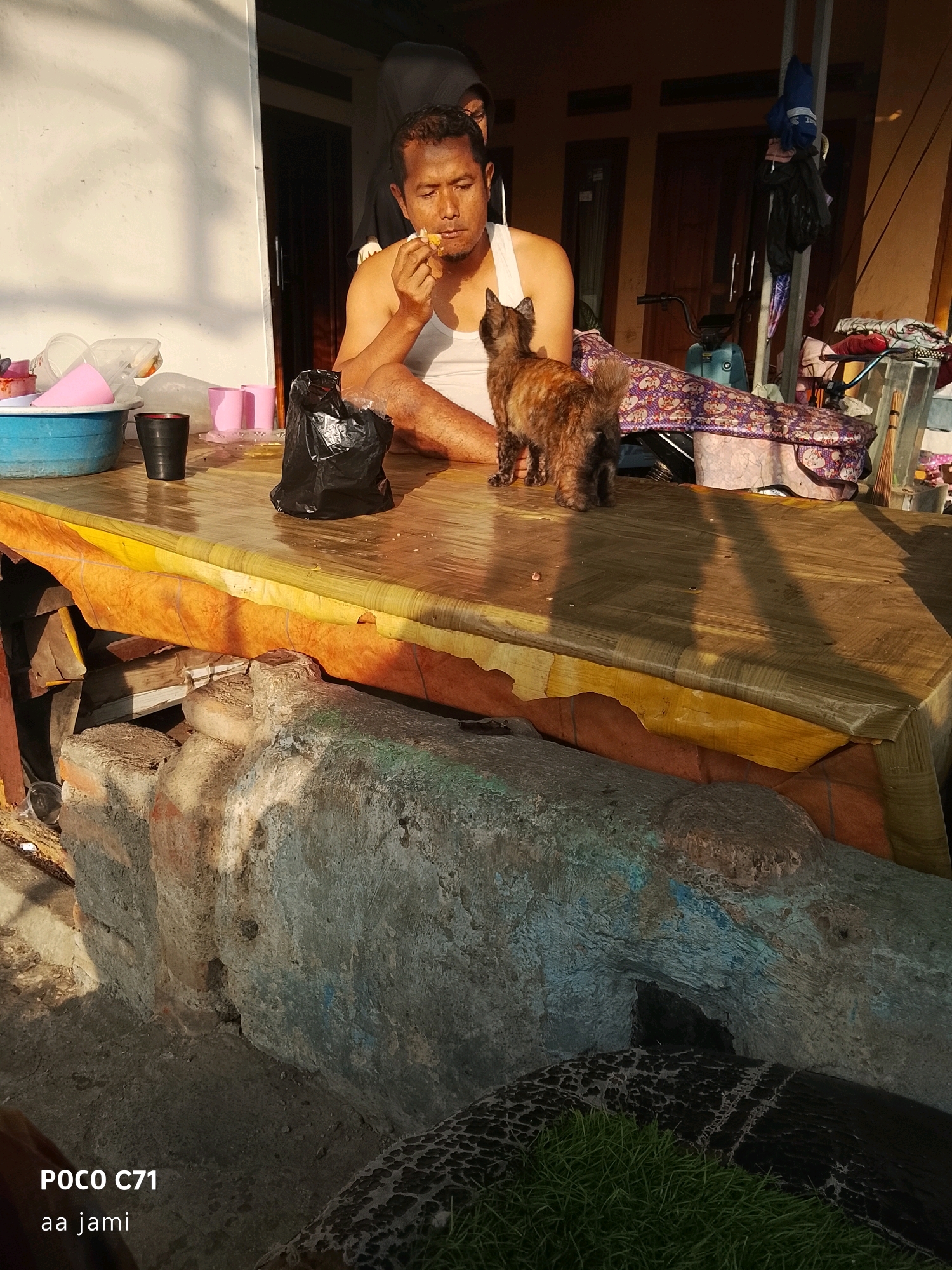 A man wearing a white tank top sits at a table, eating something. A small, dark-furred cat stands on the table in front of him, looking up at him. The table is covered with a patterned cloth, and there are various items on it, including a blue bowl, a black plastic bag, and several pink cups. In the background, a building with a white wall and wooden doors is visible. A bicycle is parked near the building, and laundry hangs on a rack. The scene appears to be outdoors, possibly in a residential area. The overall impression is one of a casual, everyday moment. There is no specific information available to determine the exact location in Tasikmalaya, Indonesia.