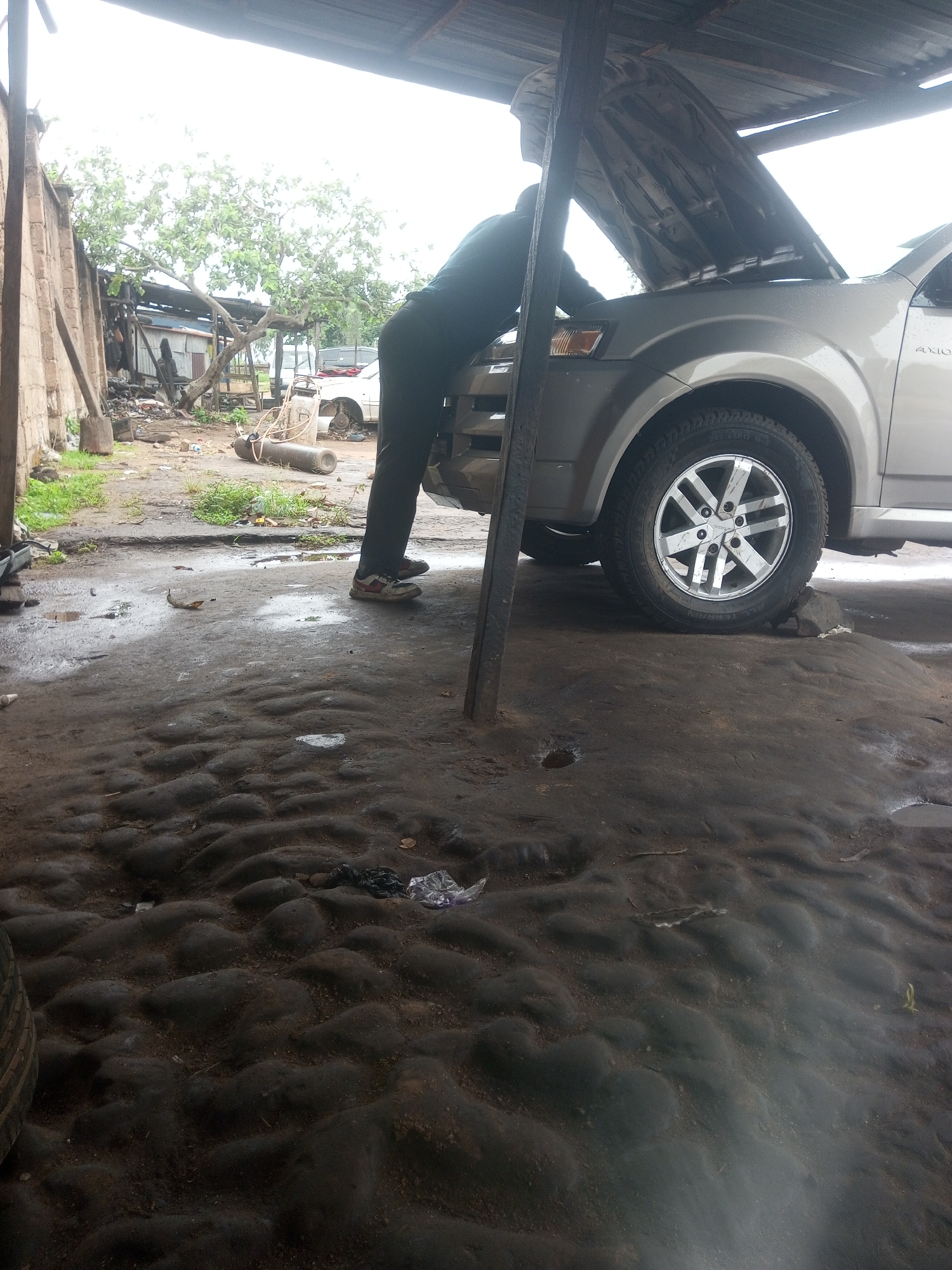 In New Stadium, Nigeria, a mechanic, wearing dark pants and a dark shirt, is working on a silver SUV with its hood open. The car is situated inside a rudimentary outdoor workshop with a corrugated metal roof supported by wooden beams. The ground is uneven, muddy, and littered with debris. To the left, a concrete wall and some wooden structures are visible, suggesting the workshop is part of a larger, unpaved area. A white sedan is parked in the background, along with a gas cylinder and various tools or car parts. The overall scene depicts a functional, albeit unrefined, automotive repair setting.
