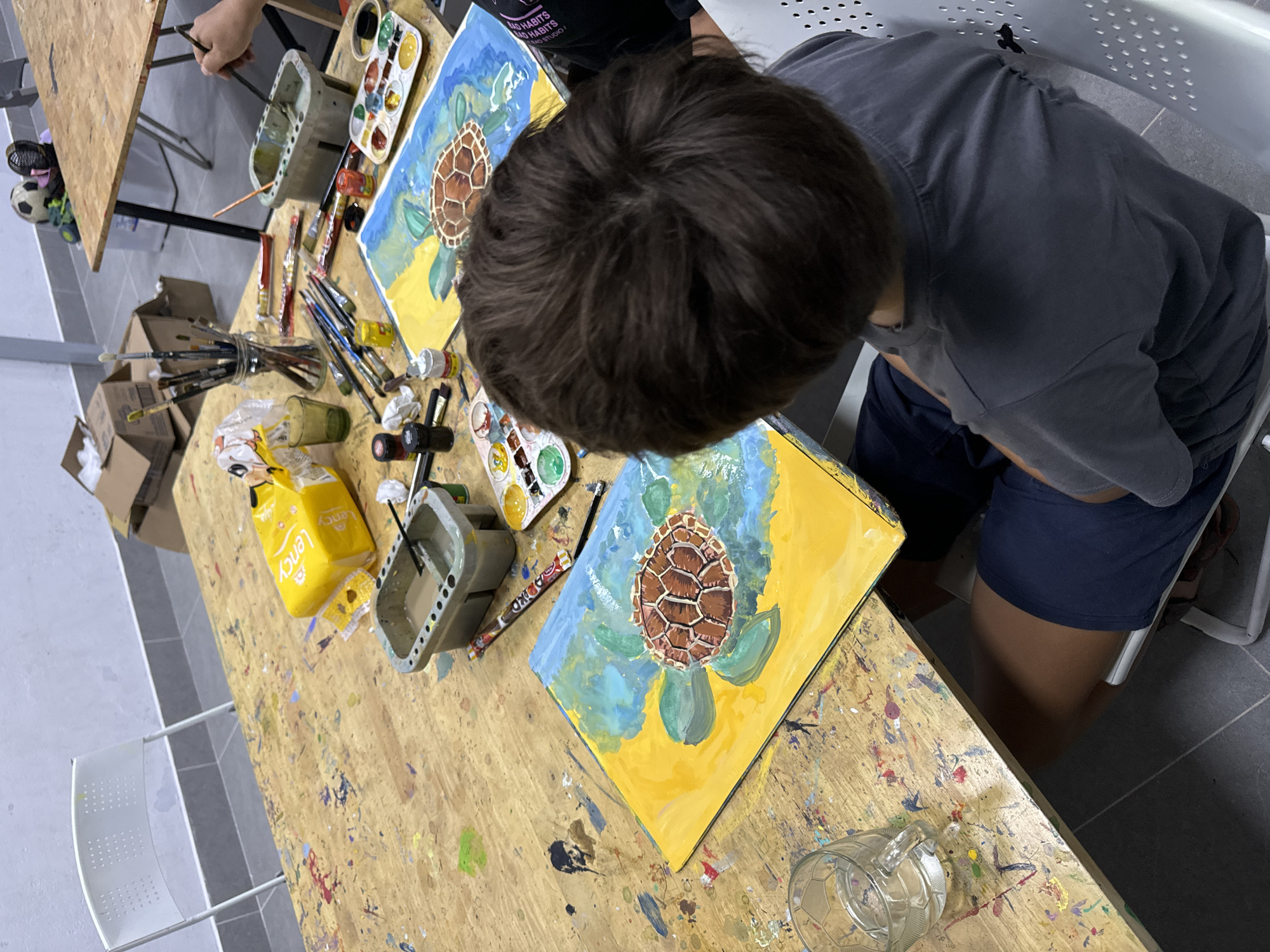 An overhead shot captures a young boy with dark hair, wearing a grey t-shirt and blue shorts, engrossed in painting a sea turtle on a wooden table. The table is cluttered with art supplies, including paintbrushes, paint palettes, jars of paint, and a glass of water. Two paintings of sea turtles are visible on the table, suggesting a shared art session. In the background, a white chair is positioned near a white wall. The overall scene suggests an art class or studio setting. The image is an indoor shot taken from a high angle, with natural light illuminating the scene. The scene does not show any indication of Mui Ne, Vietnam.