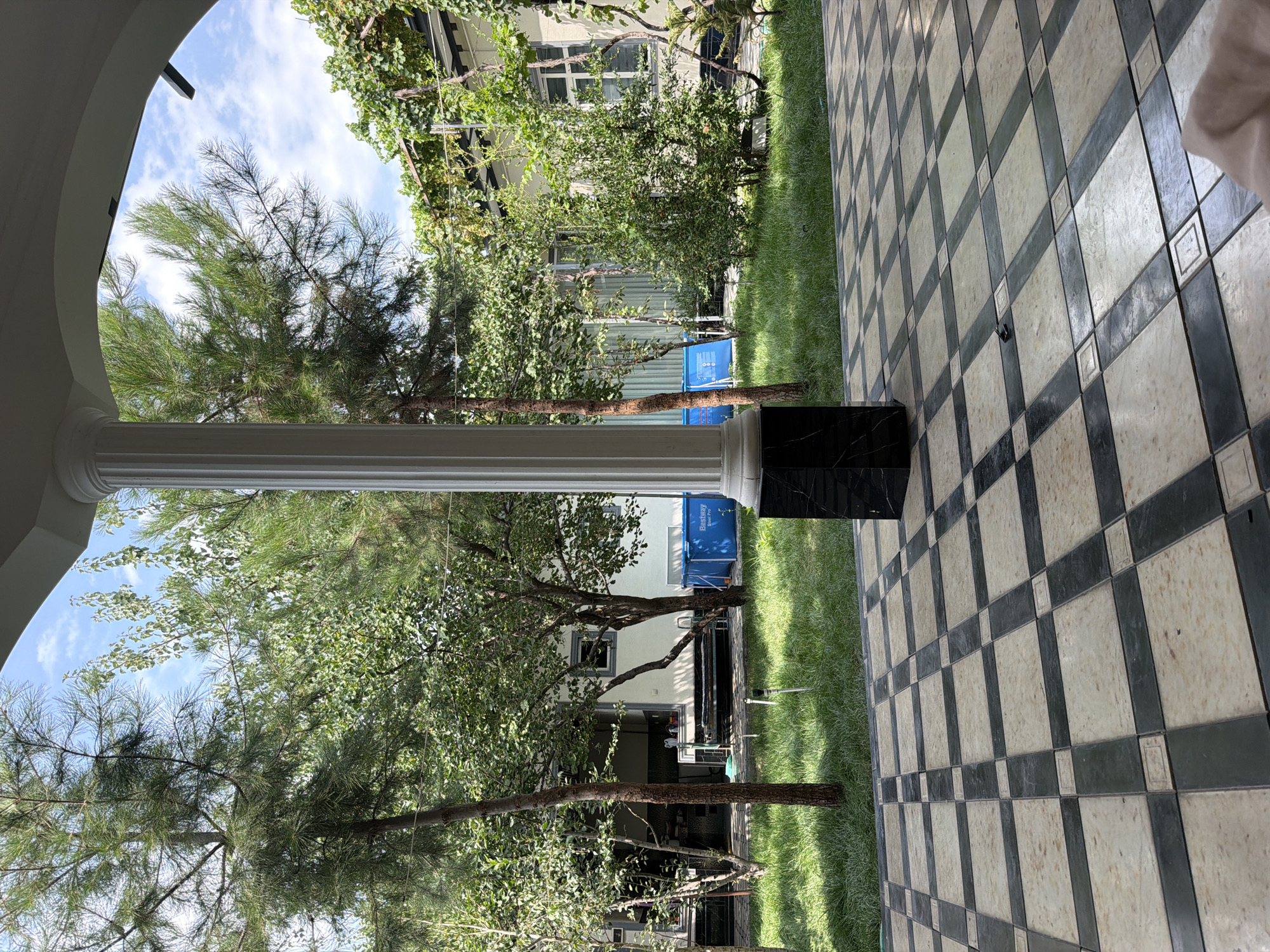 In Mindanabad, Uzbekistan, a courtyard scene unfolds. A white, fluted column stands prominently in the foreground, its base adorned with black marble. The ground is paved with a checkered pattern of light beige and dark grey tiles, with a light shadow cast on the tiles. Beyond the tiled area, a lush green lawn stretches out, dotted with various trees. A bright blue inflatable swimming pool, identified by the "Bestway Steel Pro" label, is visible in the mid-ground to the right of center. Behind the lawn and pool, a light-colored building with windows can be seen. The sky above is partly cloudy. The scene appears to be set in a residential or recreational outdoor space.