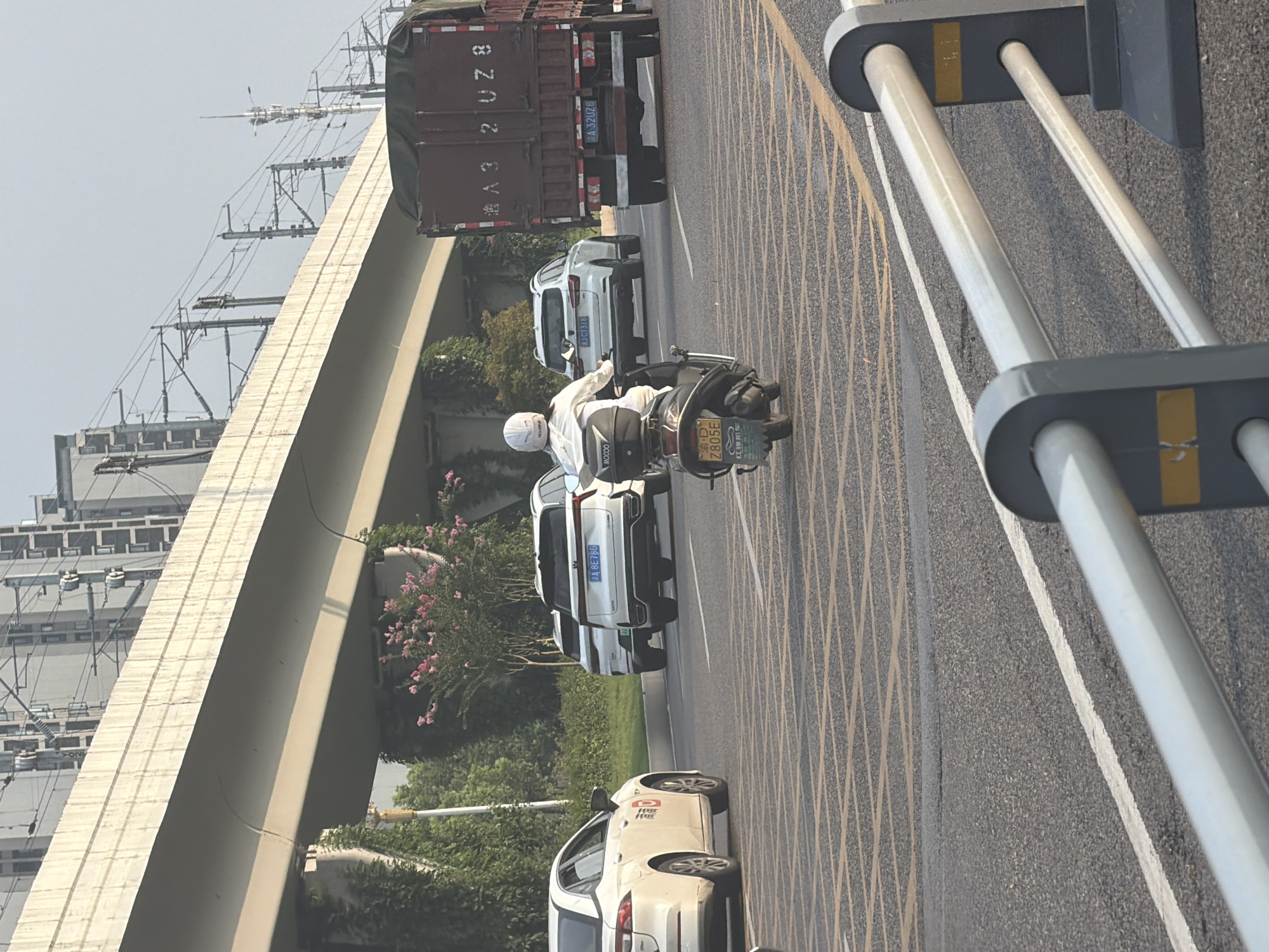 In Jiangjin, China, a multi-lane road is observed with overhead infrastructure and surrounding greenery. A white sedan is positioned on the left side of the frame, facing away from the viewer. In the center of the road, a person riding a scooter, wearing a white helmet and jacket, is moving forward. Behind the scooter, two white SUVs are in motion. To the right, a large, dark-colored truck with a tarp-covered cargo is also traveling on the road. The road surface features yellow and white lane markings, including diagonal hatching. Overhead, a concrete overpass is visible with electrical wires and poles. The background consists of buildings and trees, suggesting an urban environment.