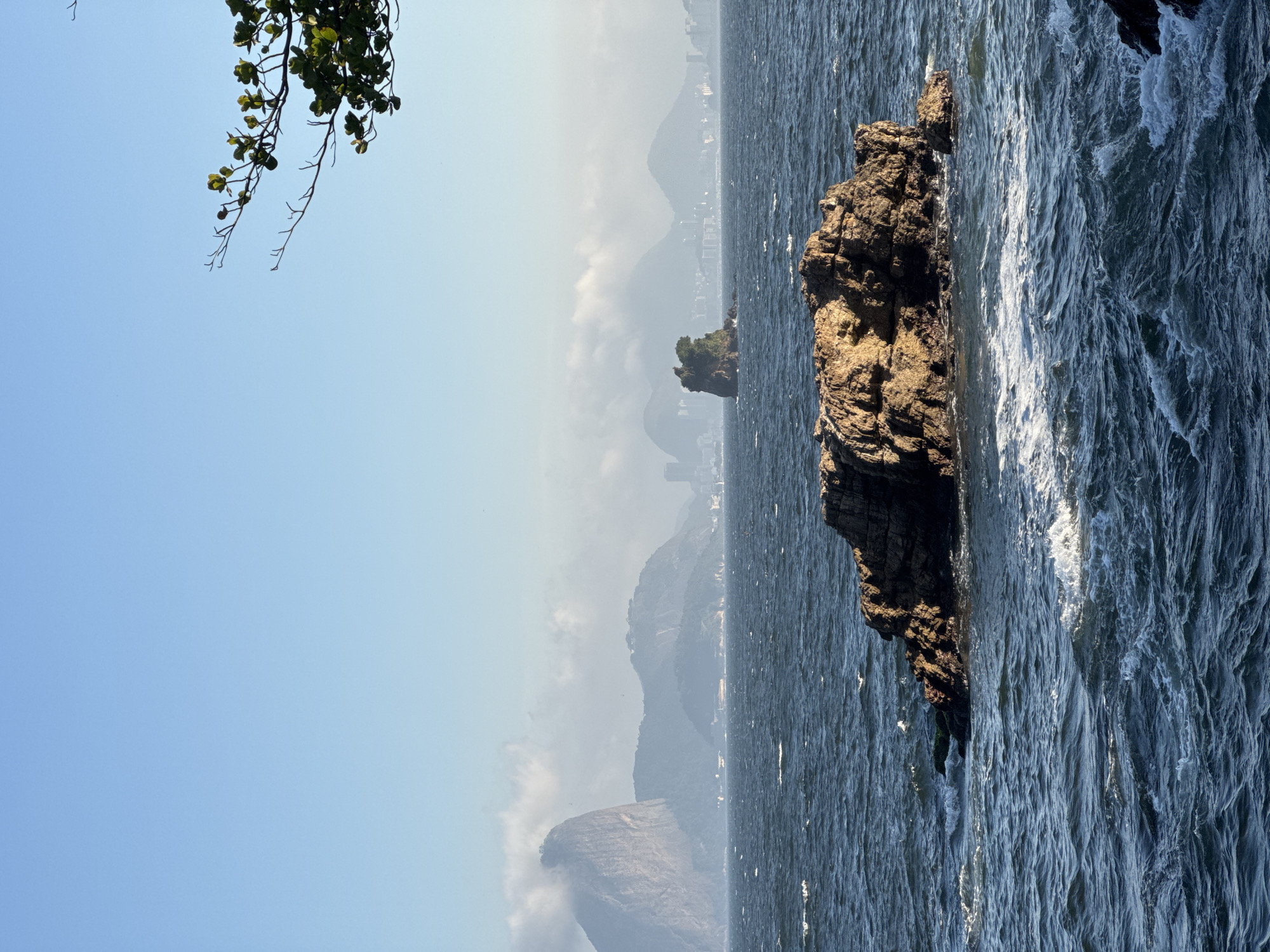 In the city of Niterói, Brazil, a rugged, sun-drenched rock formation dominates the foreground, emerging from the dark, choppy waters of the ocean. The waves crash against the rock, creating white foam that contrasts with the deep blue of the sea. In the mid-ground, a smaller, tree-topped island sits in the ocean, further out from the shore. In the background, a hazy skyline of mountains and buildings stretches across the horizon under a clear blue sky. The iconic Sugarloaf Mountain, shrouded in mist, is visible on the left, a recognizable landmark of the nearby city of Rio de Janeiro. A tree branch with green leaves extends into the frame from the top right corner, adding a touch of nature to the scenic view. There are no people or animals visible in the image.