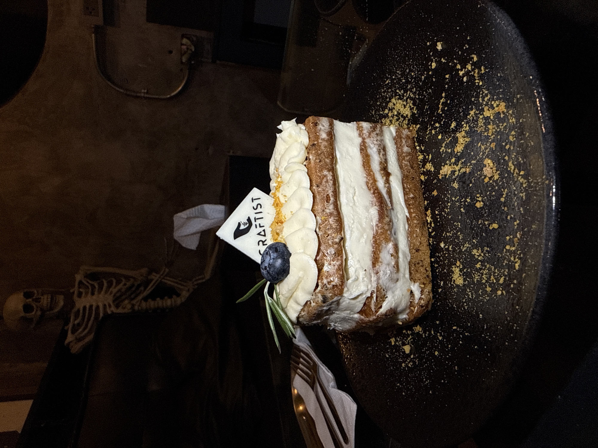 A slice of layered cake with white frosting, a blueberry, and rosemary sprig is presented on a dark plate, sprinkled with crumbs. A small flag with a logo is stuck into the top of the cake. A fork and knife are visible to the left of the cake, resting on a napkin. In the background, a decorative skeleton is visible leaning against a dark surface. The lighting is dim, suggesting an indoor environment, possibly a restaurant or cafe. There is no explicit indication of the location being in Phuket City Municipality, Thailand; this information is provided as context.