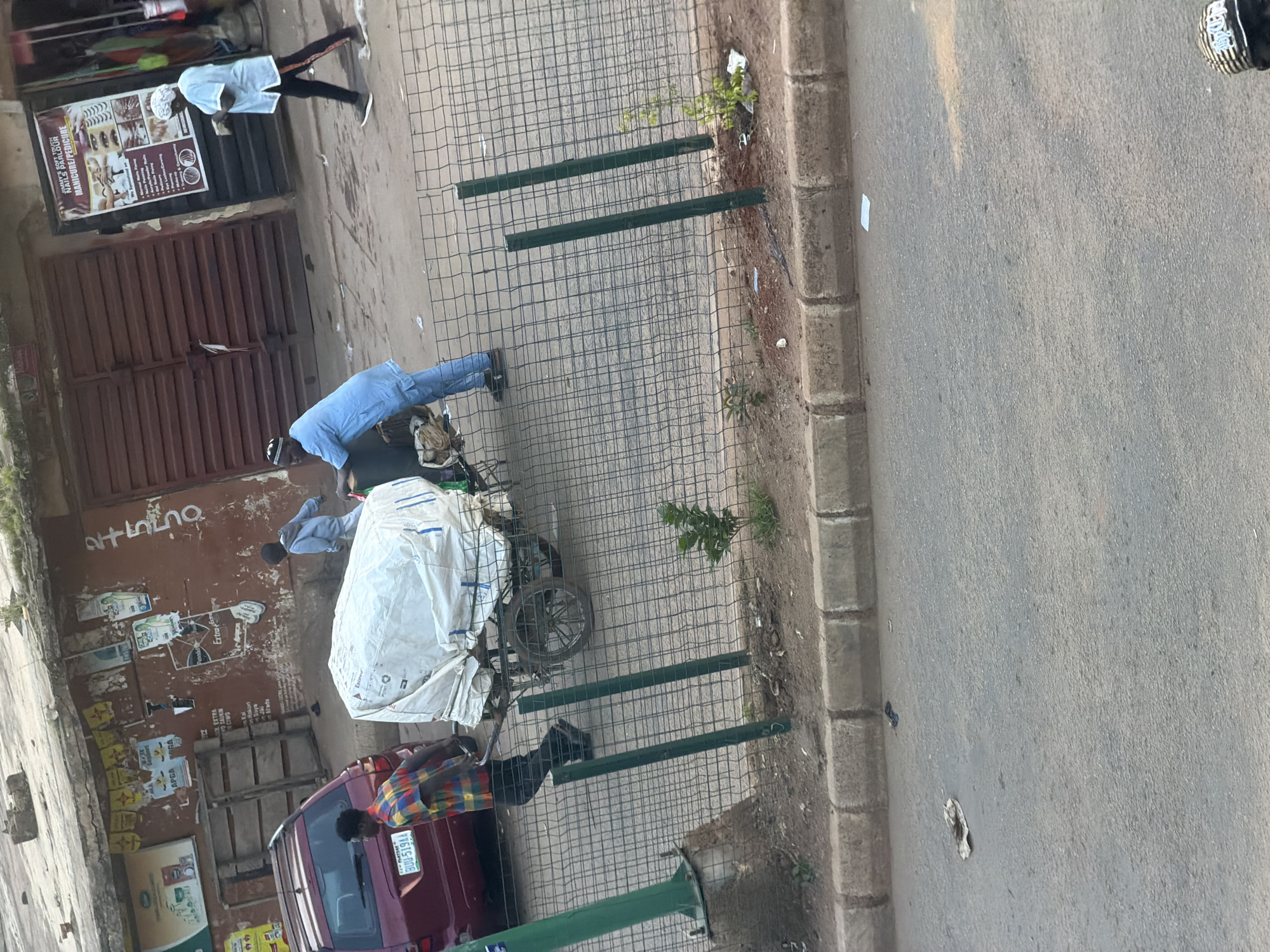 This media file captures an outdoor scene in Bukuru, Nigeria, likely during daylight hours due to the shadows cast. The setting appears to be a street or sidewalk adjacent to a building with a large, weathered brown door and a sign for "NAILS PARLOUR MANICURE/PEDICURE." Various posters and advertisements are affixed to the building's facade.
In the foreground, a man wearing a blue shirt and pants is bending over a bicycle laden with a large, white tarp-covered object. He appears to be adjusting or securing the load. Another person, dressed in a white shirt and head covering, is walking away from the building, their back to the camera. Further in the background, a person in a colorful checkered shirt is seen near a maroon van with a license plate reading "BUU-519AA."
The scene is bordered by a grid of metal fencing, with some green painted posts interspersed. Small plants and weeds are growing in the dirt and gravel along the edge of the paved area. The overall impression is one of daily life and commerce in an urban or semi-urban Nigerian environment. The lighting suggests it might be late afternoon, with the sun casting long shadows.