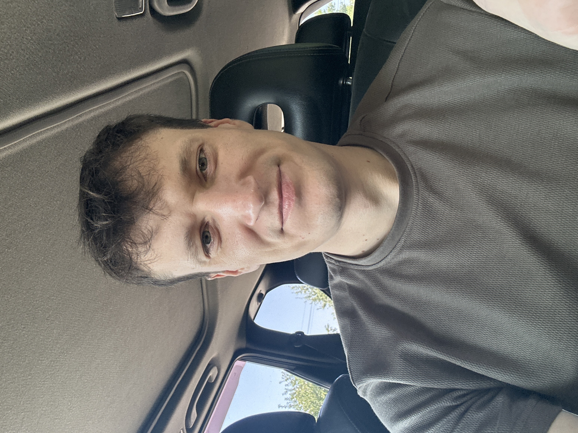 A medium shot selfie shows a man inside a car. The man has fair skin, short brown hair, and is looking directly at the camera with a slight smile. He is wearing a grey t-shirt. The interior of the car is visible, including the headrest of a black leather seat, part of the car's roof lining, and the edge of the sunroof. Sunlight is visible through the windows, and some greenery can be seen outside. The image is likely taken in Makiv, Ukraine, as suggested by the prompt.