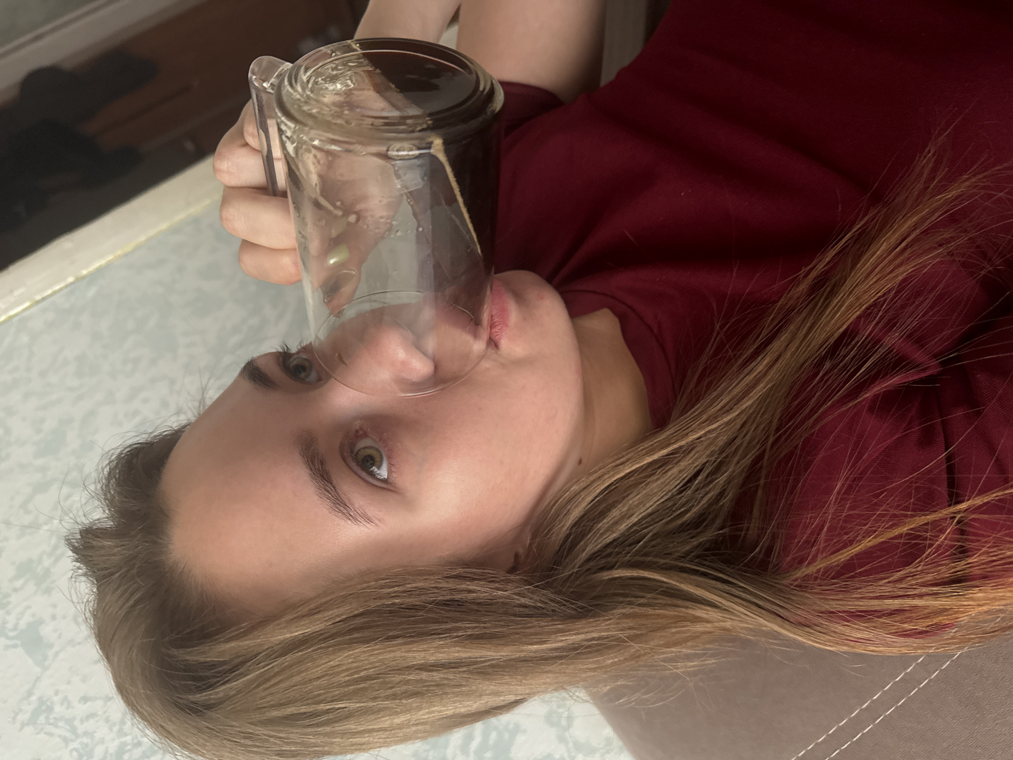 A young woman with long, light brown hair drinks from a glass jar. She is wearing a dark red t-shirt. The background appears to be an indoor setting with a patterned wallpaper and furniture. There is no indication of the specific location in Chaikovsky, Russia.