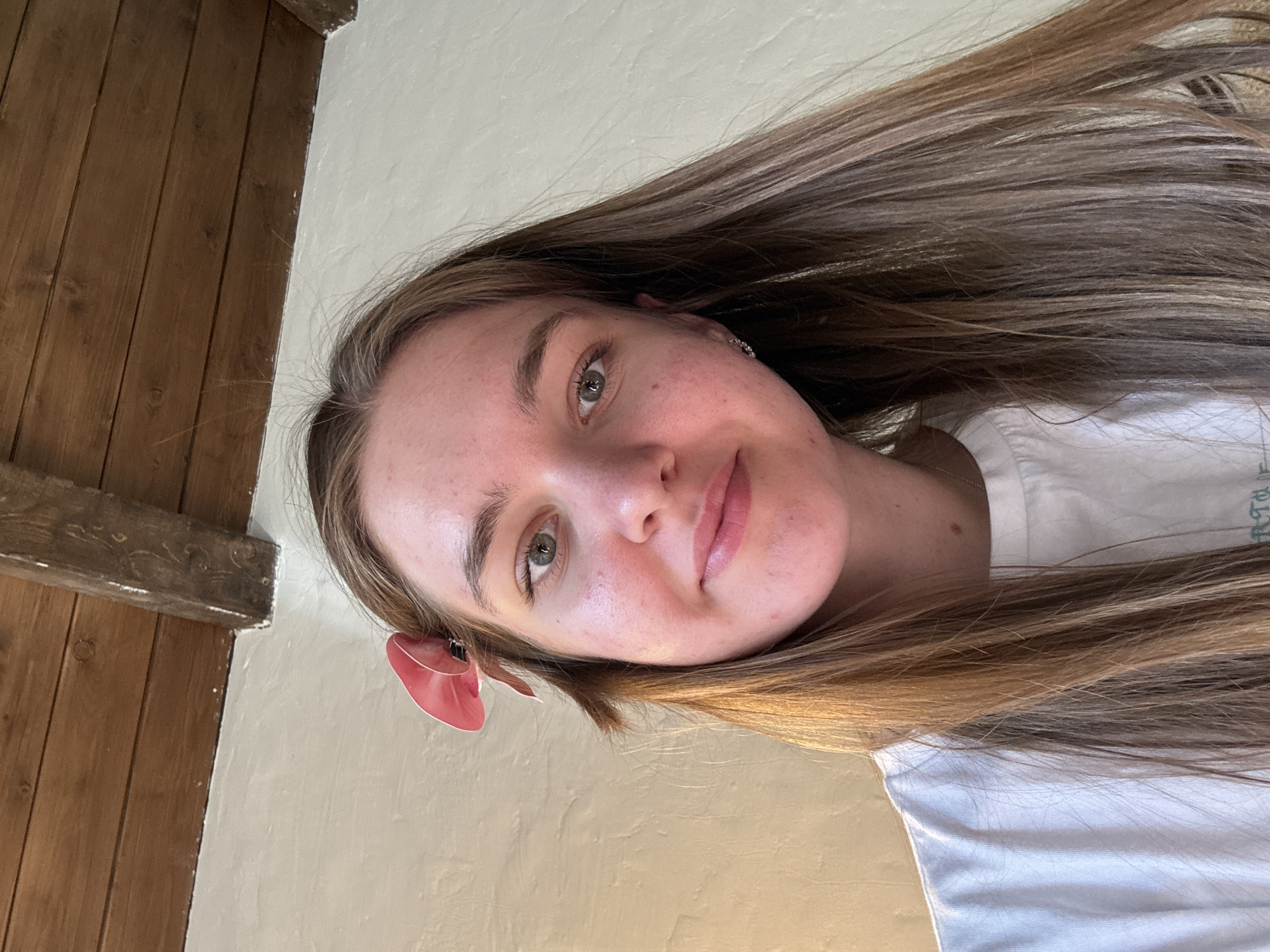 A young woman with long, light brown hair is shown in a close-up selfie. She has fair skin, light eyes, and is wearing a white t-shirt. A decorative red accessory is visible near her ear. The background includes a light-colored wall and wooden paneling, suggesting an interior setting. The image provides no information to identify the location in Sibir', Russia.